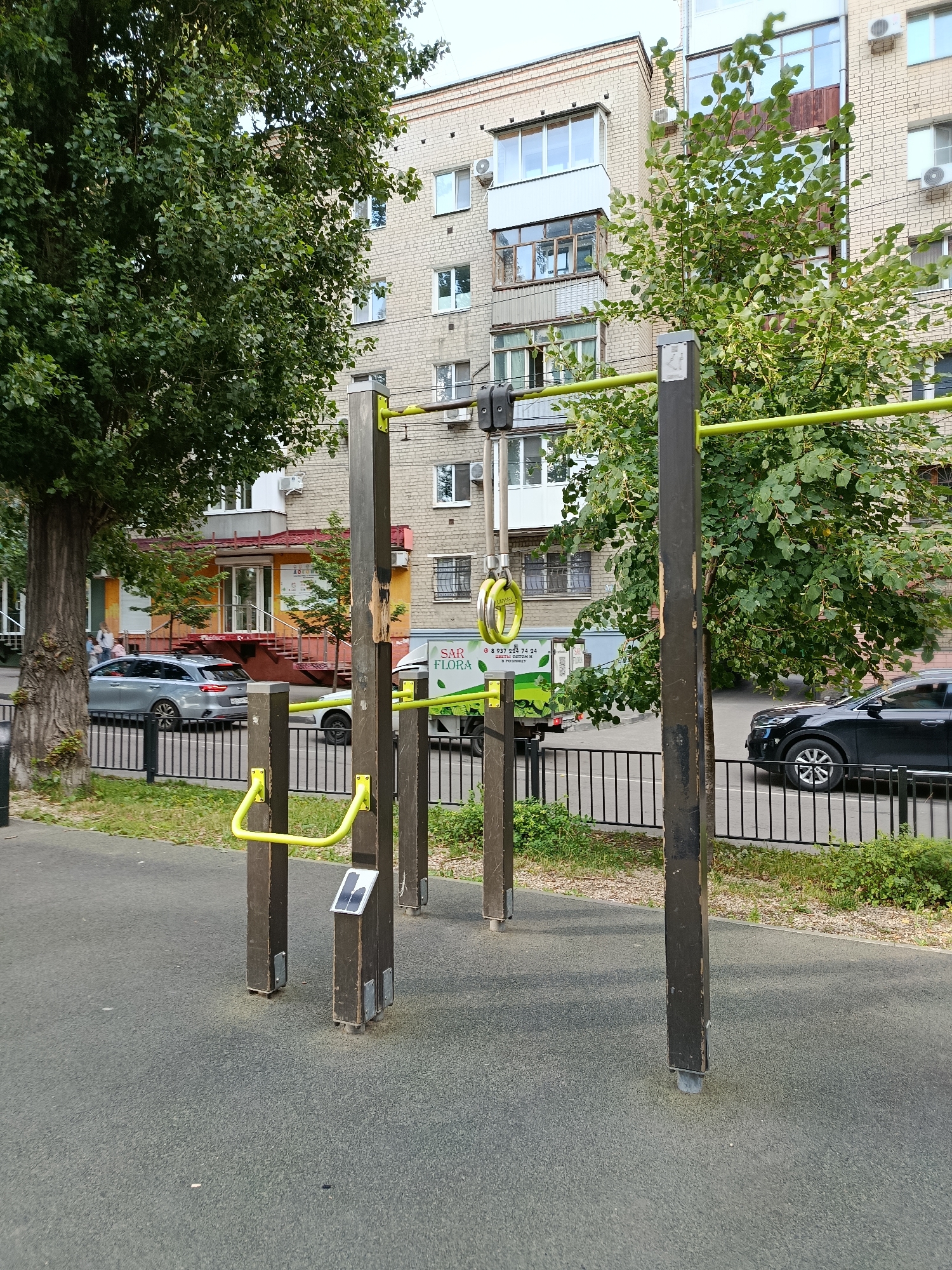 In an outdoor urban setting, an exercise apparatus with wooden posts and a bright yellow horizontal bar is situated on a dark grey rubberized surface. Attached to the apparatus are yellow handles and a pair of gymnastic rings. Behind the apparatus, a black metal fence delineates a street where a silver station wagon and a black SUV are parked. Across the street, a light brick apartment building with multiple windows and balconies stands, partially obscured by leafy green trees. A commercial vehicle with the text "SAR FLORA" is also visible. The scene suggests a public space in a city like Saratov, Russia, likely a park or a residential courtyard.