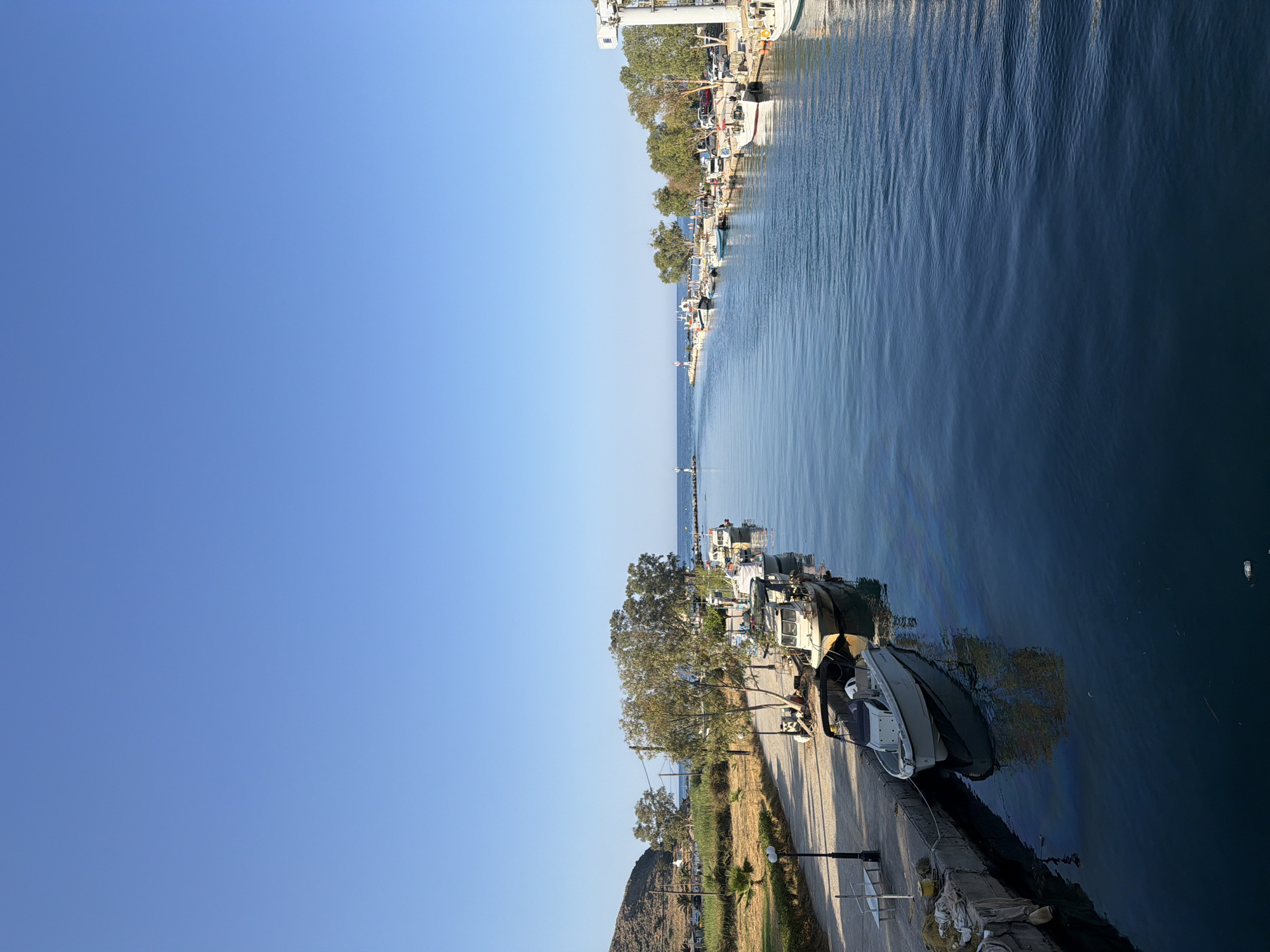 This is a vertical, eye-level, medium shot of a canal in Georgioupoli, Greece, under a clear blue sky. The canal is lined with various boats docked on both sides. On the left, a paved walkway runs alongside the canal, with trees and vegetation behind it. In the distance, a hill is visible. On the right, more boats are docked, and a white structure, possibly a crane, stands near the end of the canal, where it meets the sea. A lighthouse is visible in the distance, marking the entrance to the canal from the sea. The water in the canal is calm and reflects the sky and the surrounding trees. There are no people visible in the image.