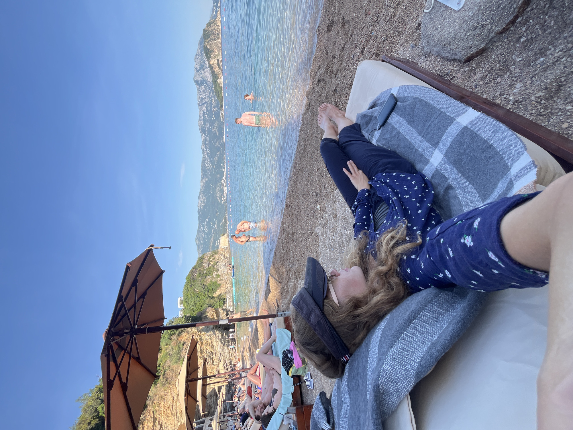 A woman in a blue floral dress and black pants is lying on a beach lounge chair with a gray and white plaid blanket. She is wearing a visor. In the background is the coastline of Budva, Montenegro, with mountains and clear water where people are swimming. Other beachgoers are visible under beach umbrellas.