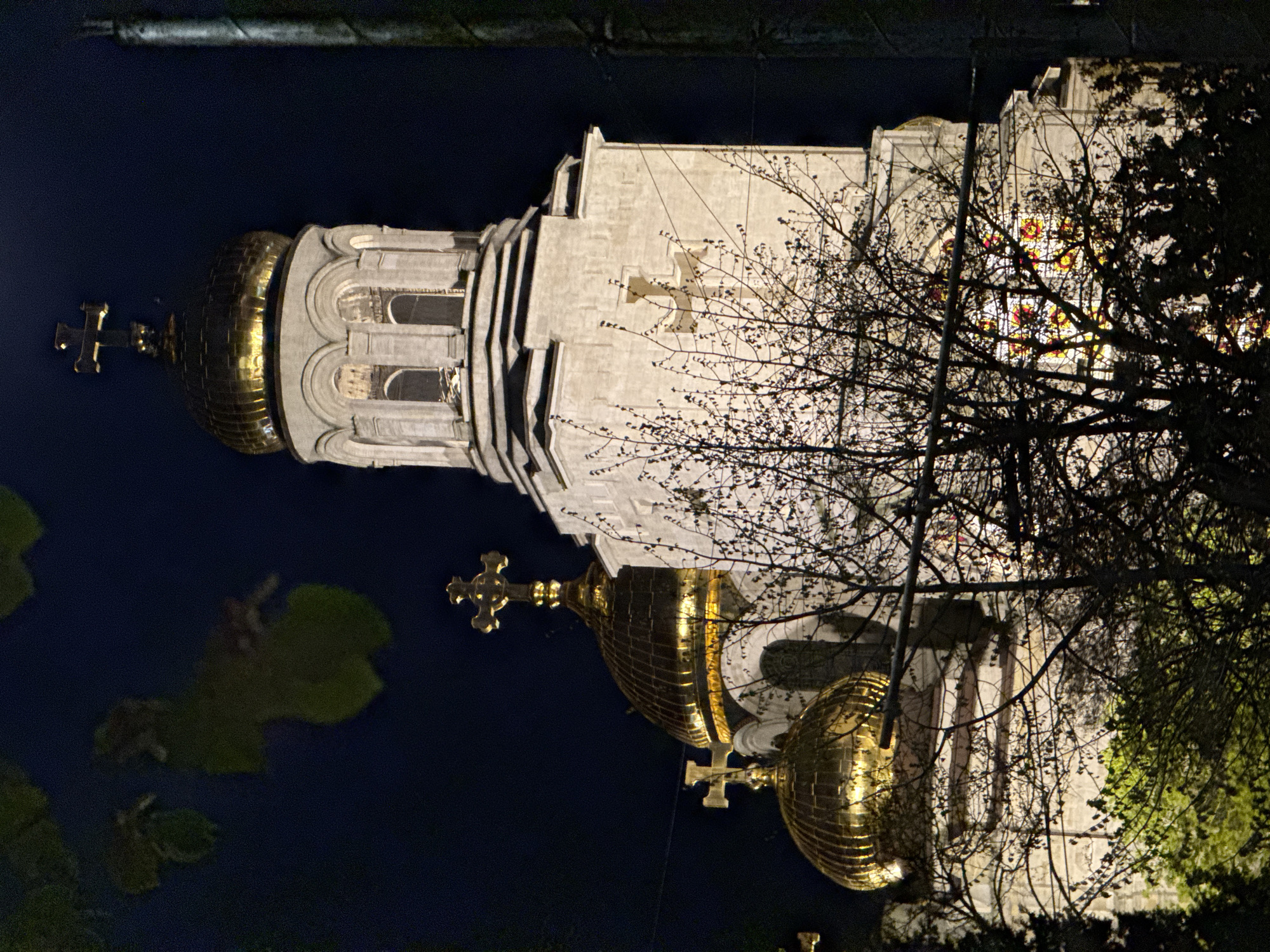 The image displays a grand Eastern Orthodox church, likely in Varna, Bulgaria, illuminated at night. The impressive building features a light-colored, possibly stone or stucco, facade against a dark night sky. Multiple golden onion domes, each crowned with a distinct Orthodox cross, are prominent. Two large, shimmering golden domes are visible, with the leftmost one featuring a textured, almost brick-like pattern that reflects the artificial light.
The architecture is rich with detail, including arched windows and decorative elements on the main tower. A large, carved cross is embedded in the central part of the church's wall. Towards the right, vibrant stained glass windows, glowing with patterns of red and yellow, are visible through a dense network of bare tree branches. Some sparse green foliage is also present on the edges, suggesting surrounding trees or shrubs.
The scene is tranquil and devoid of people or activity, focusing entirely on the majestic structure under artificial illumination. The lighting emphasizes the architectural intricacies and the golden brilliance of the domes, creating a striking nocturnal view of this significant religious landmark. No visible text is present in the image.