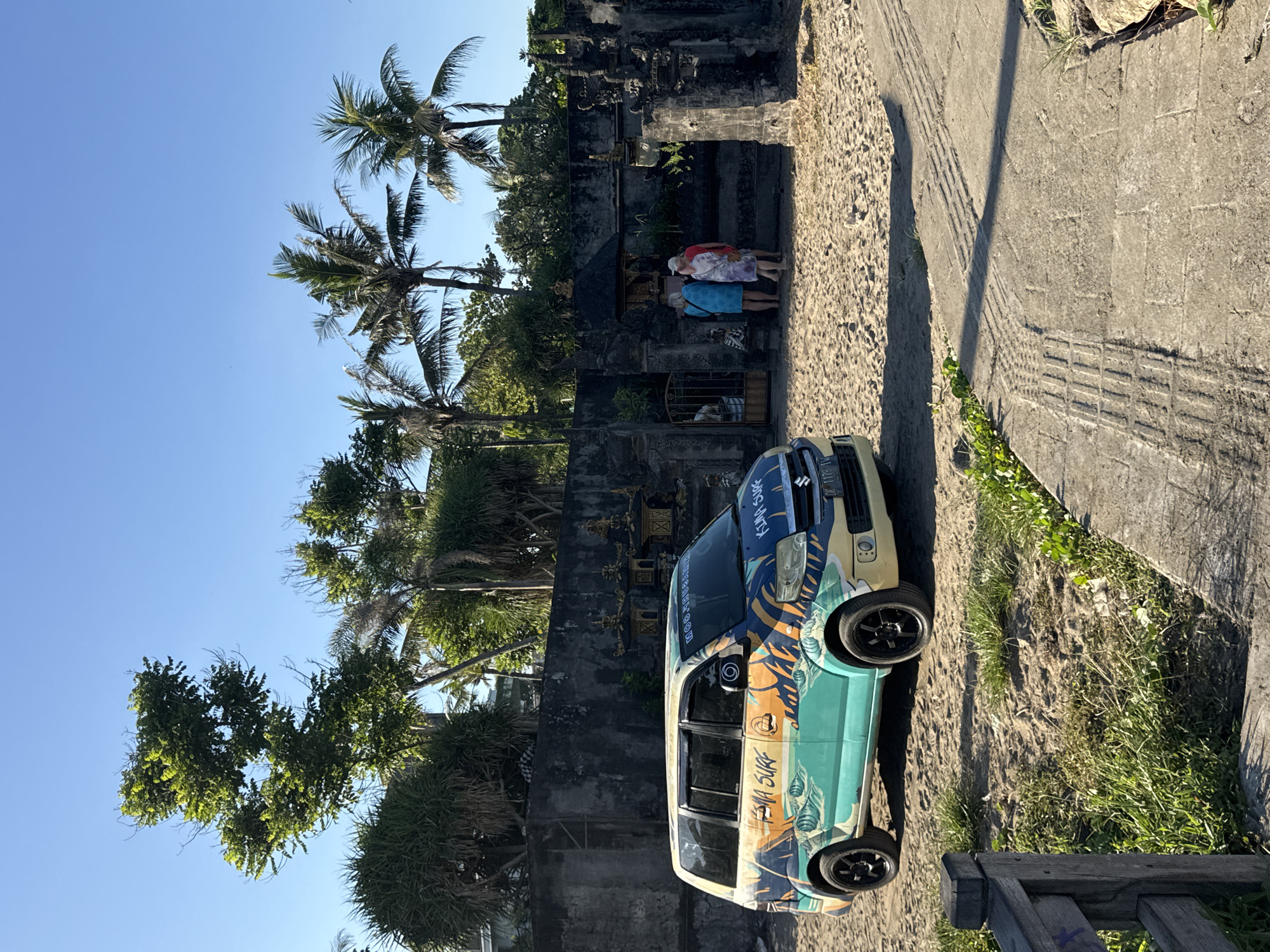 Captured in Kuta, Indonesia, the image shows a parked Suzuki decorated with a surf-themed graphic, situated on a sandy patch adjacent to a paved sidewalk. Behind the car stands a traditional Balinese structure of dark stone, where two people are standing and looking in. Palm trees are seen overhead. The sky is clear and blue.