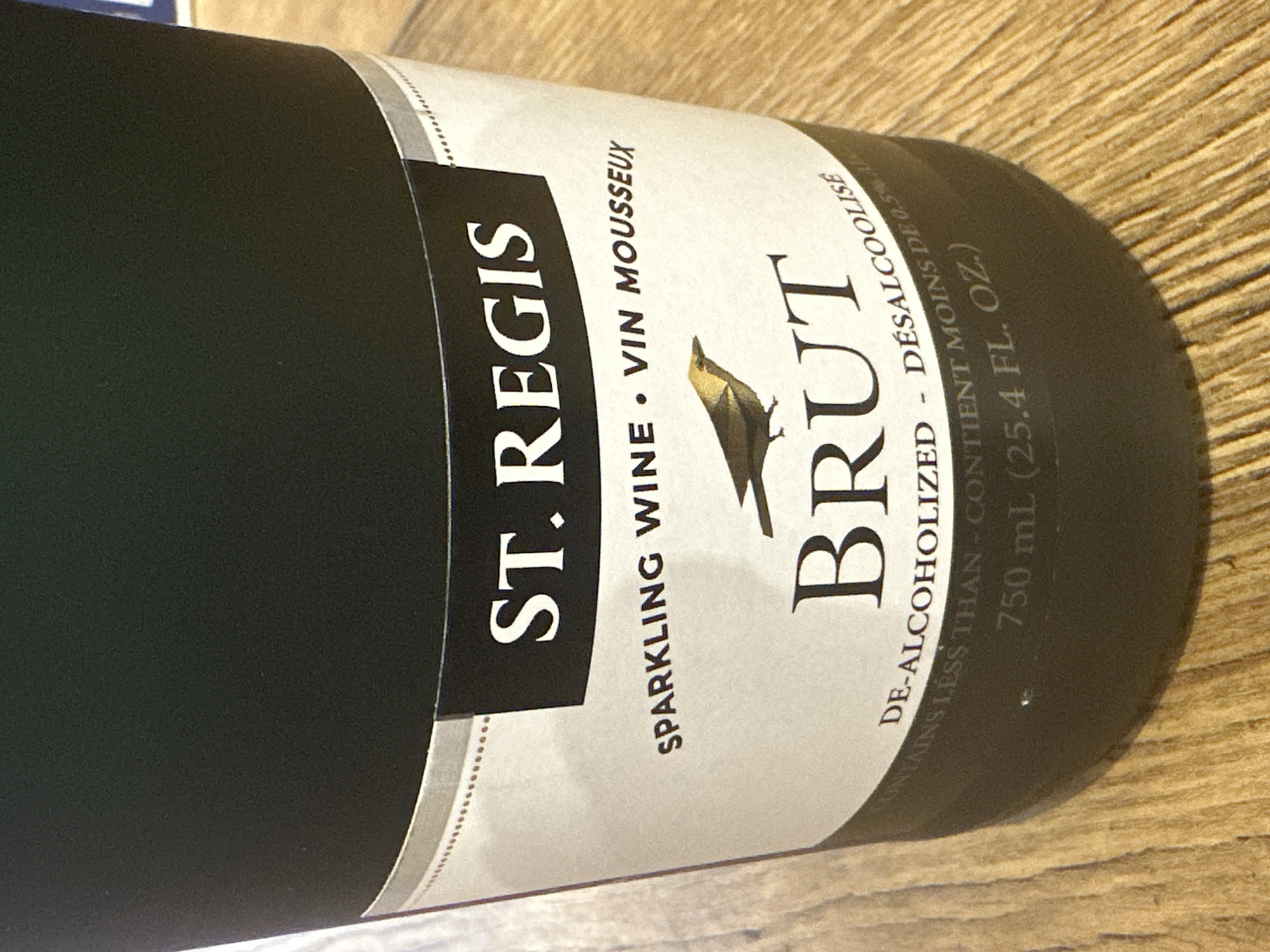 A close-up, eye-level, indoor shot shows a dark gray bottle of ST. REGIS Sparkling Wine, Brut. The label is white with black text and a small brown bird illustration. The text reads "ST. REGIS", "SPARKLING WINE • VIN MOUSSEUX", "BRUT", "DE-ALCOHOLIZED - DÉSALCOOLISE", and "CONTAINS LESS THAN - CONTIENT MOINS DE 0.5% ALC. BY VOL.", "750 mL (25.4 FL. OZ.)". The bottle is resting on a light brown wooden surface, possibly a table. The background is a dark, out-of-focus area, suggesting an indoor setting. There are no people visible in the image. The scene appears to be in Camp Hill, United States of America, as per the prompt.