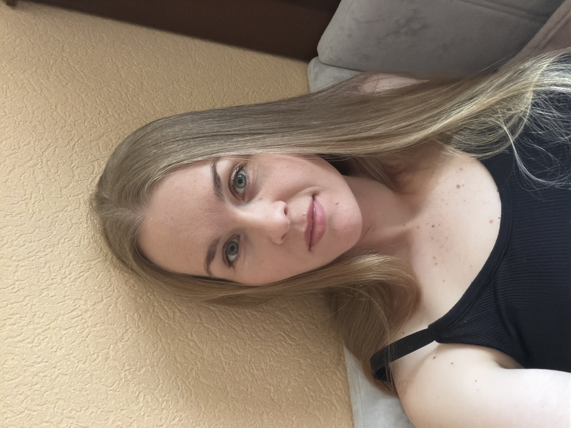 This is a selfie of a woman with long, blonde hair and blue eyes, wearing a black tank top. She is sitting on a couch in front of a textured, yellow wall. The lighting is natural and bright, suggesting it is daytime. There is a hint of a wooden object or furniture to the right of the frame. The background is slightly blurred, with the focus on the woman's face and upper body. The image appears to be taken indoors. The setting does not provide any specific information to locate it in Khabarovsk, Russia.