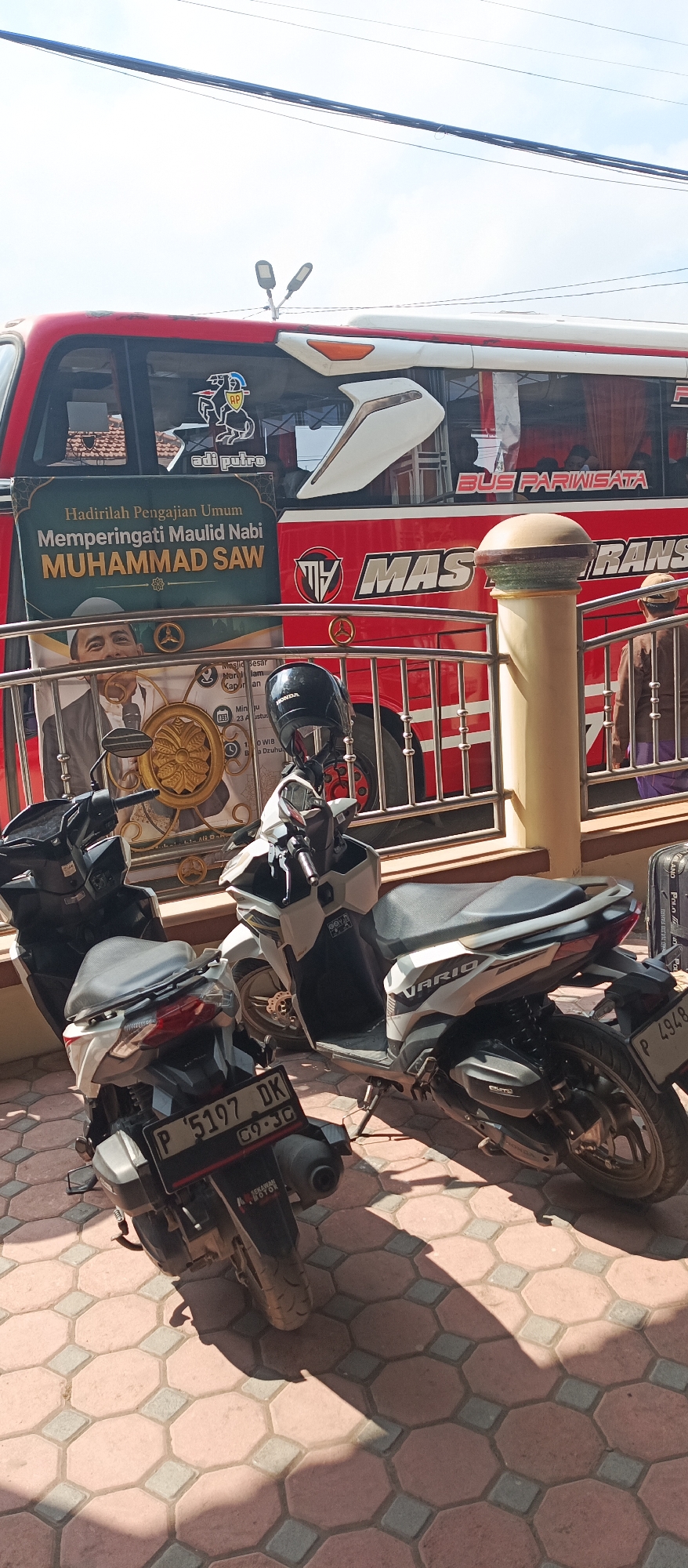Two motorcycles are parked on a patterned pavement in front of a red and white bus. The bus has "BUS PARIWISATA" written on its side and a banner that reads "Hadirlah Pengajian Umum Memperingati Maulid Nabi MUHAMMAD SAW" which translates to "Attend the General Recitation Commemorating the Birthday of the Prophet MUHAMMAD SAW." There are also images of a man and a gold circular emblem on the banner. One motorcycle is black and white, the other is white. A black helmet rests on the handlebars of the white motorcycle. A person's back is visible on the right side of the image, wearing brown clothing and a head covering. The scene appears to be outdoors under a bright sky with power lines visible overhead. The license plate of the black and white motorcycle reads "P 5197 DK". The location is identified as Kapongan, Indonesia.