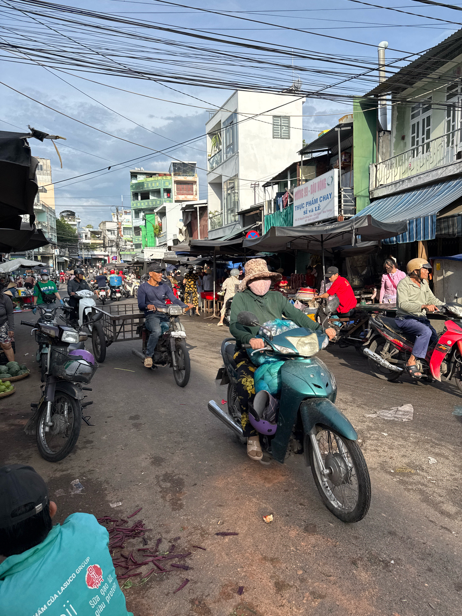 The image captures a bustling street scene in Nha Trang, Vietnam, likely a market or busy thoroughfare. The sky is overcast with a mix of clouds and patches of blue. Overhead, a dense network of electrical wires crisscrosses the sky, a common sight in Vietnamese cities. Buildings lining the street are a mix of styles and colors, some with balconies and awnings, suggesting a mix of residential and commercial use.
The street is populated with numerous motorbikes, the primary mode of transportation. People are riding them, some wearing helmets and face coverings, indicating protection from the sun or pollution. A few individuals are walking, and a child is visible in the foreground.
Objects scattered on the street include baskets of produce, possibly fruits and vegetables, and what appear to be cut pieces of purple vegetables, like eggplant or okra. In the middle ground, a motorbike with a trailer is being ridden by a man. Another person on a motorbike is seen in the foreground, wearing a wide-brimmed hat and a face covering. The road surface is asphalt, showing signs of wear and some water stains. The overall impression is one of daily life in a vibrant urban environment.
