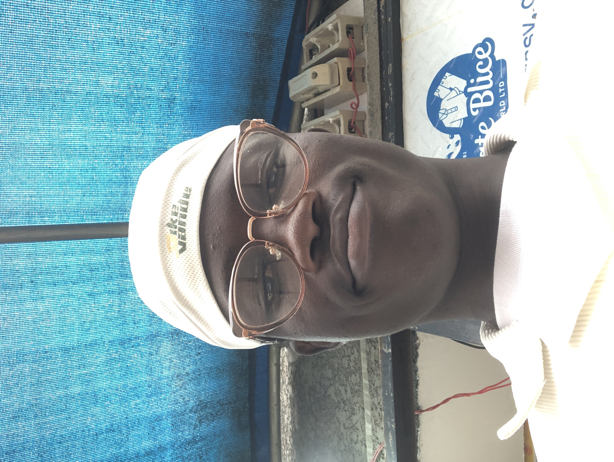 In the city of Ado, Nigeria, a young man is captured in a close-up, selfie-style photograph. He is wearing a white cap and clear-rimmed glasses that partially obscure his eyes. His facial expression is a gentle smile, with his mouth closed. The background consists of a vibrant blue textured surface, possibly a shade cloth or awning, common in outdoor settings in Nigeria. Below this, a textured grey wall or surface is visible, on which three electrical fuse holders with red wires are mounted. To the right of the frame, a portion of a sign featuring a logo with shirts and the text "te Blice RLD LTD" is visible. The man is wearing a light-colored, possibly cream or off-white, collared shirt, evident from the ribbed neckline. The lighting suggests an outdoor environment, with sunlight illuminating the scene and casting subtle shadows. The overall impression is an informal, candid shot taken during the day.
