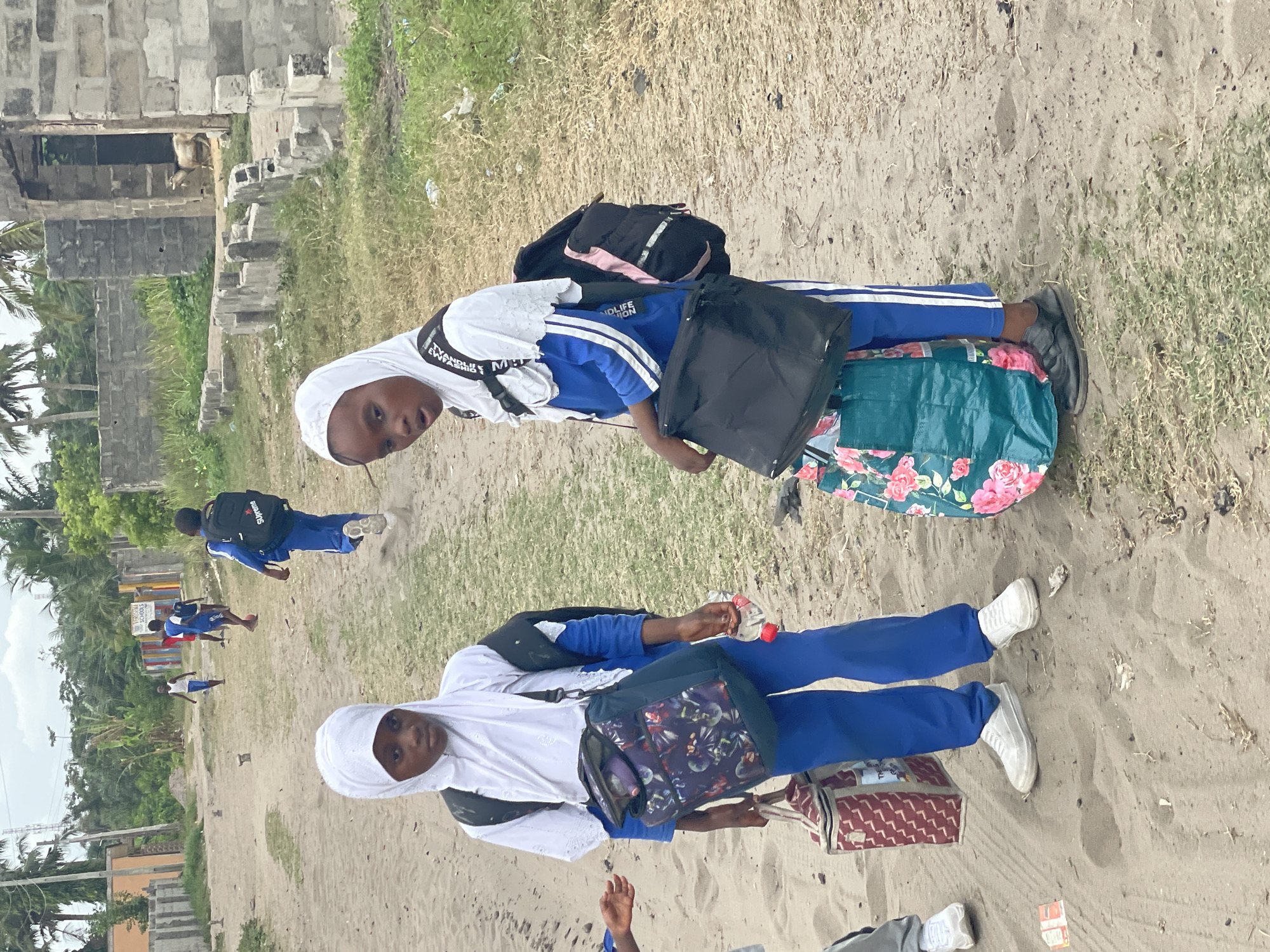 Two schoolgirls in blue and white uniforms, wearing white hijabs, stand on a sandy area in Badagry, Nigeria. One girl carries a black backpack and a floral bag, while the other has a patterned shoulder bag and a patterned tote. Several other students in similar uniforms are walking away in the background. Buildings and greenery are visible in the background, as is some trash. The location appears to be near an urban area.
