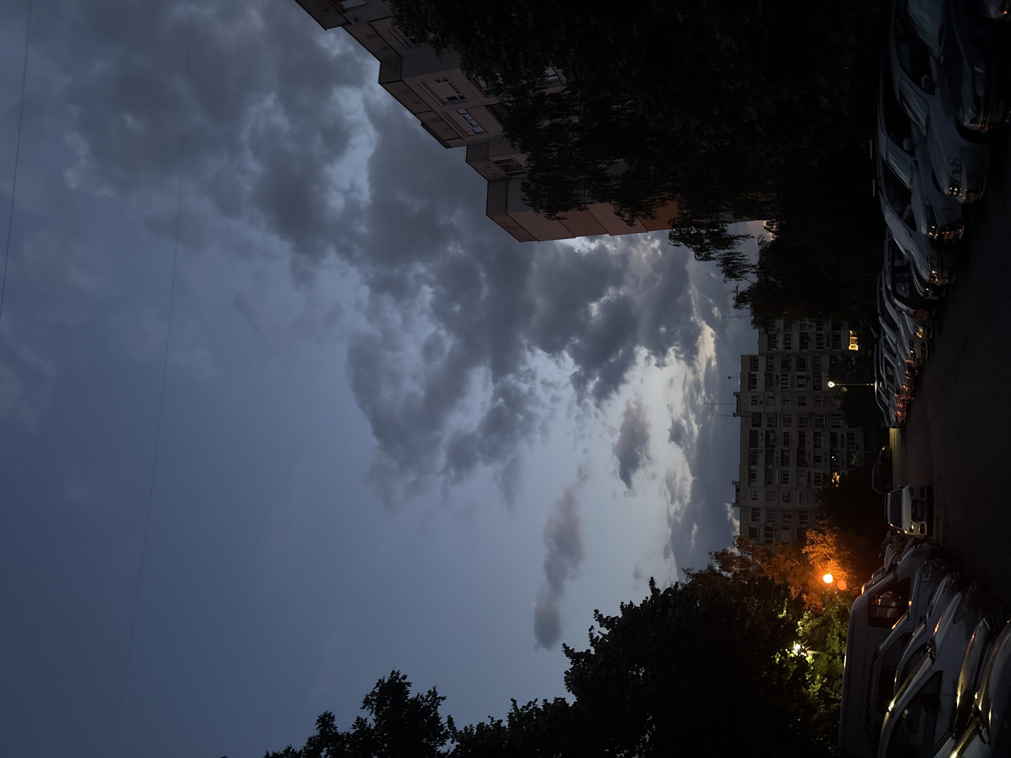 A nighttime shot captures an urban scene. A parking lot filled with numerous cars is visible in the foreground, with a white sedan positioned centrally. Tall apartment buildings, characteristic of Soviet-era architecture, line the background. Dark trees frame the scene on the left and right. A streetlamp with an orange glow illuminates the left side of the image. The sky is dark and cloudy, with a thin wire stretched across it.