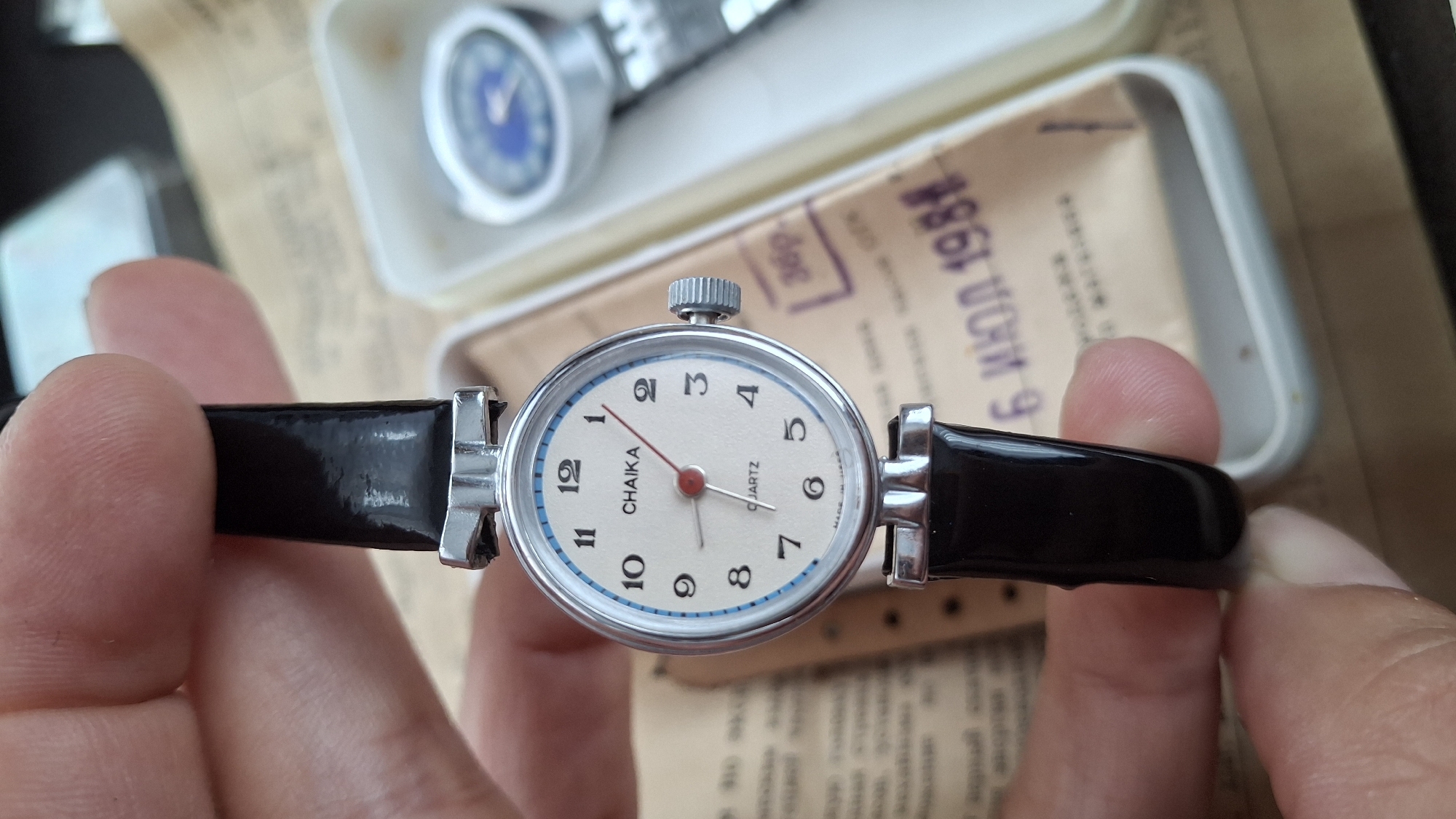 A close-up, eye-level shot shows a person's hand holding a silver wristwatch with a black strap. The watch face is cream-colored with black numerals, a red second hand, and the brand name "CHAIKA" above the word "QUARTZ." The watch is positioned over a document with Cyrillic text and the date "6 ИЮЛ 1981". In the background, out of focus, is a white box containing another watch with a blue face. The overall environment appears to be indoors, possibly on a table, with soft lighting. The image does not contain any information that places it in the city of Kempelevo, Russia.