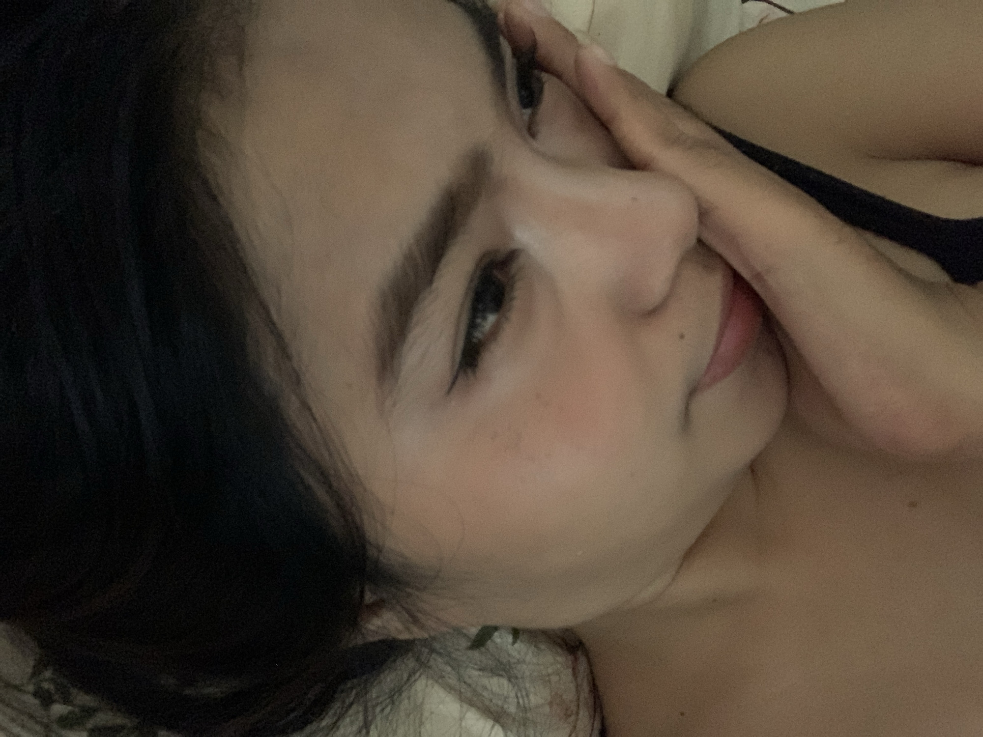 A close-up, angled portrait shot features a woman lying down, possibly in a bed, with soft, dim lighting suggesting evening or night. Her face, in profile, is partially obscured by her hand resting against her cheek and nose. Her dark hair falls around her. She appears to be looking slightly away from the camera, and her expression seems contemplative or serene. Visible elements include her eyes, eyebrows, nose, mouth, and a hint of her shoulder and a dark strap of clothing, possibly a tank top or bra. The background is indistinct, consisting of light-colored bedding or pillows. No specific location cues are visible beyond an indoor setting. There is no visible text.