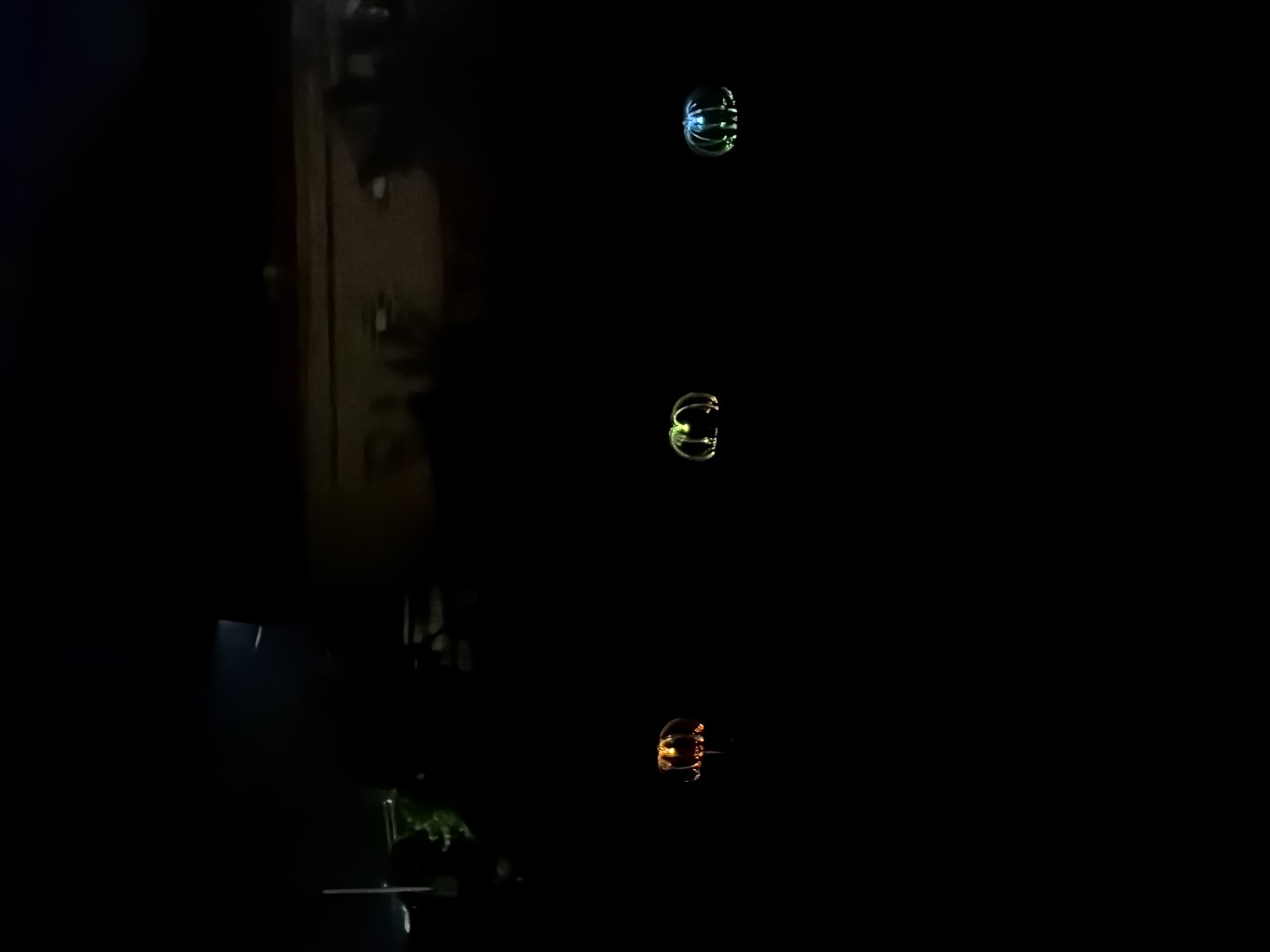 This nocturnal image depicts an outdoor scene in the city of Akkuzovo, Russia. Three distinct, illuminated decorative objects are positioned in the foreground on an indistinct dark ground. From left to right, the first object emits an orange-yellow light, appearing as a carved pumpkin or lantern with visible facial features. The second object glows greenish-yellow, and the third object glows blue-green; both exhibit a rounded, rib-like structure, suggesting decorative lanterns or stylized pumpkins. The background is predominantly dark and out of focus. Faint architectural outlines, consistent with buildings or structures, are vaguely discernible in the upper mid-ground, along with indistinct dark shapes suggestive of distant foliage or landscaping. A subtle, linear light streak is visible in the upper left portion of the frame, and a faint light source appears on the far right edge of the background. No individuals or discernible actions are present.