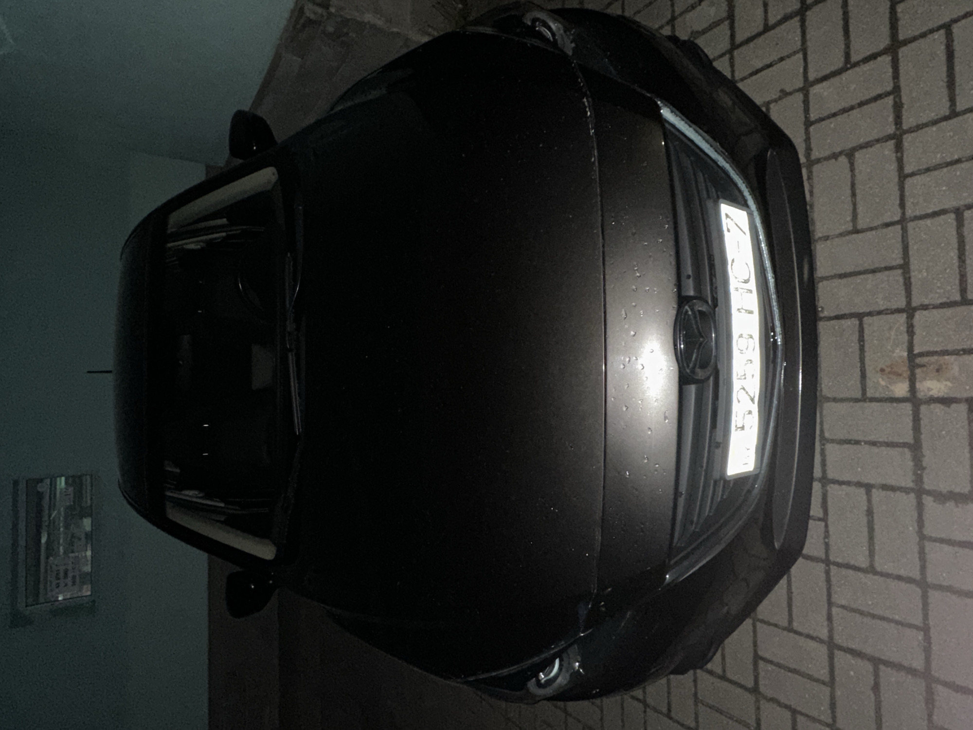 The image shows a dark-colored car parked on a paved surface, likely a parking lot or driveway. The vehicle's front end is prominently visible, with the license plate displaying "5253 HC-7". The Mazda logo is visible on the car's front grill. The background suggests an urban environment with a wall and window visible on the left side of the frame. The scene appears to be in Minsk, Belarus, based on the license plate format. The lighting conditions suggest either dusk or nighttime, with artificial lighting present.