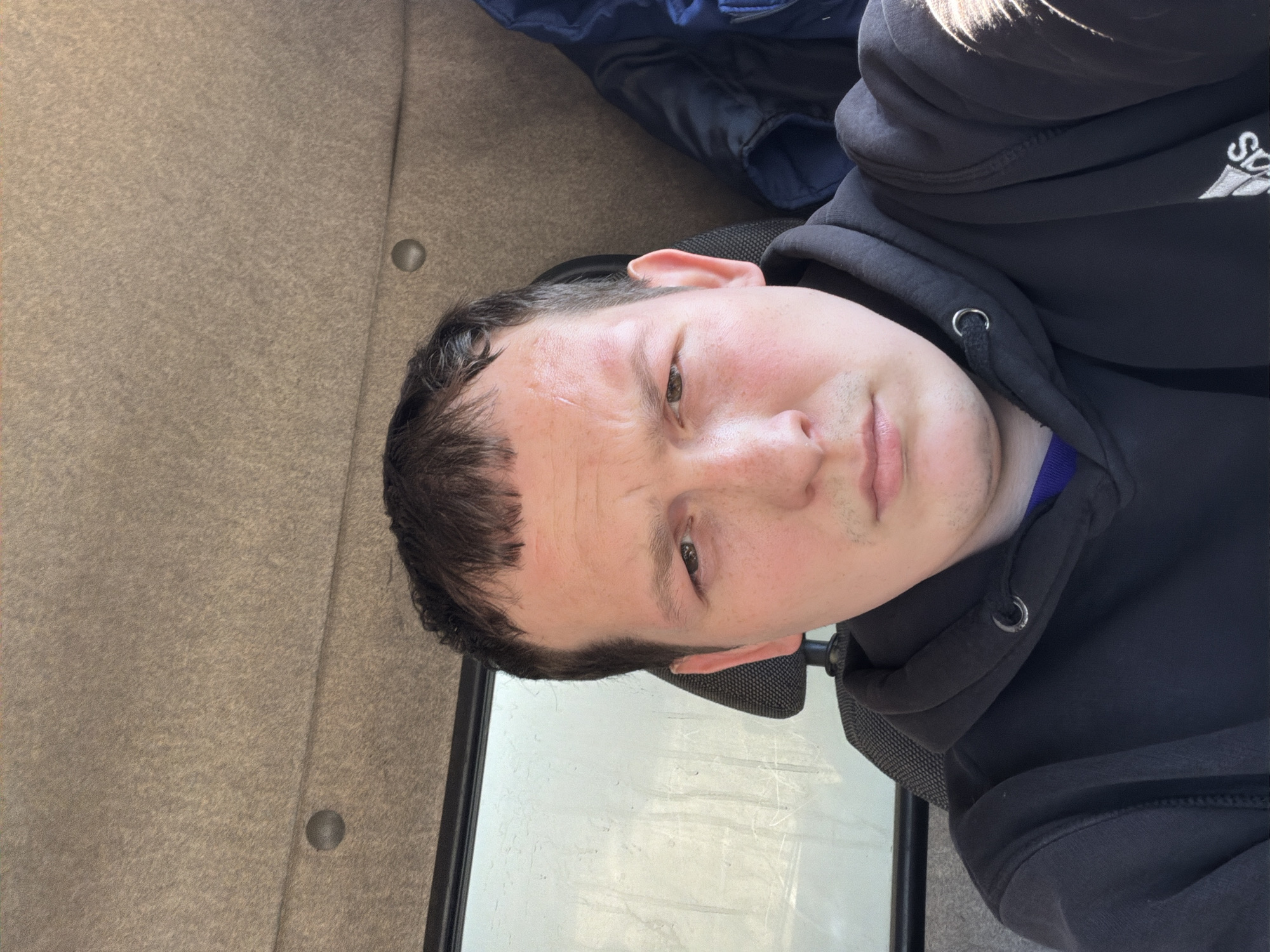 A close-up, rotated image of a man with short, dark hair and light skin is shown. He is wearing a black hoodie with a logo partially visible on the upper right side. The hood is up. A blue garment is visible near the top of the frame, possibly a jacket. The man has a small scrape on his forehead. He is looking at the camera with a neutral expression. A window with a white surface is behind him. To the left, a surface with button is visible. The location within Chaikovsky, Russia cannot be determined from this image.