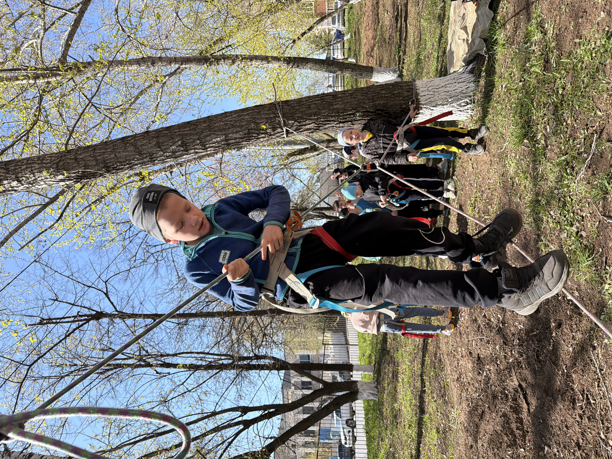 The image captures a scene in what appears to be a park-like setting in Izhevsk, Russia, evident from the building architecture and tree species commonly found in the region. A young boy, equipped with climbing gear including a harness and helmet, is suspended mid-air, secured by ropes that are attached to nearby trees. He is holding onto one of the ropes, seemingly participating in a rope-course activity. Other people, including both adults and children, are also present in the background, some appearing to be waiting their turn, suggesting a group outing or organized event. The location is characterized by trees with budding leaves, indicating spring or early summer. The ground is a mix of grass and bare earth.