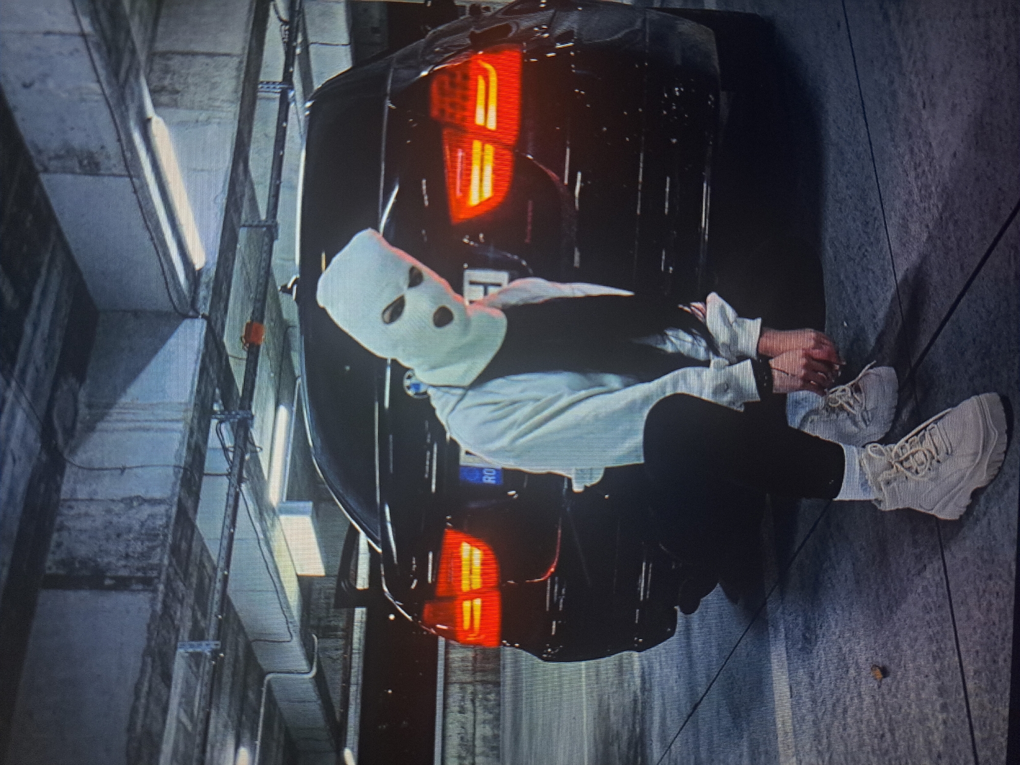 A person wearing a white ski mask and light-colored clothing sits with their hands bound behind their back against the rear of a black car with illuminated tail lights. The car’s license plate is partially visible. The setting appears to be a dimly lit parking garage with exposed concrete walls, pipes, and overhead lighting fixtures. There are no indications in the image suggesting this location is in Yerevan, Armenia.