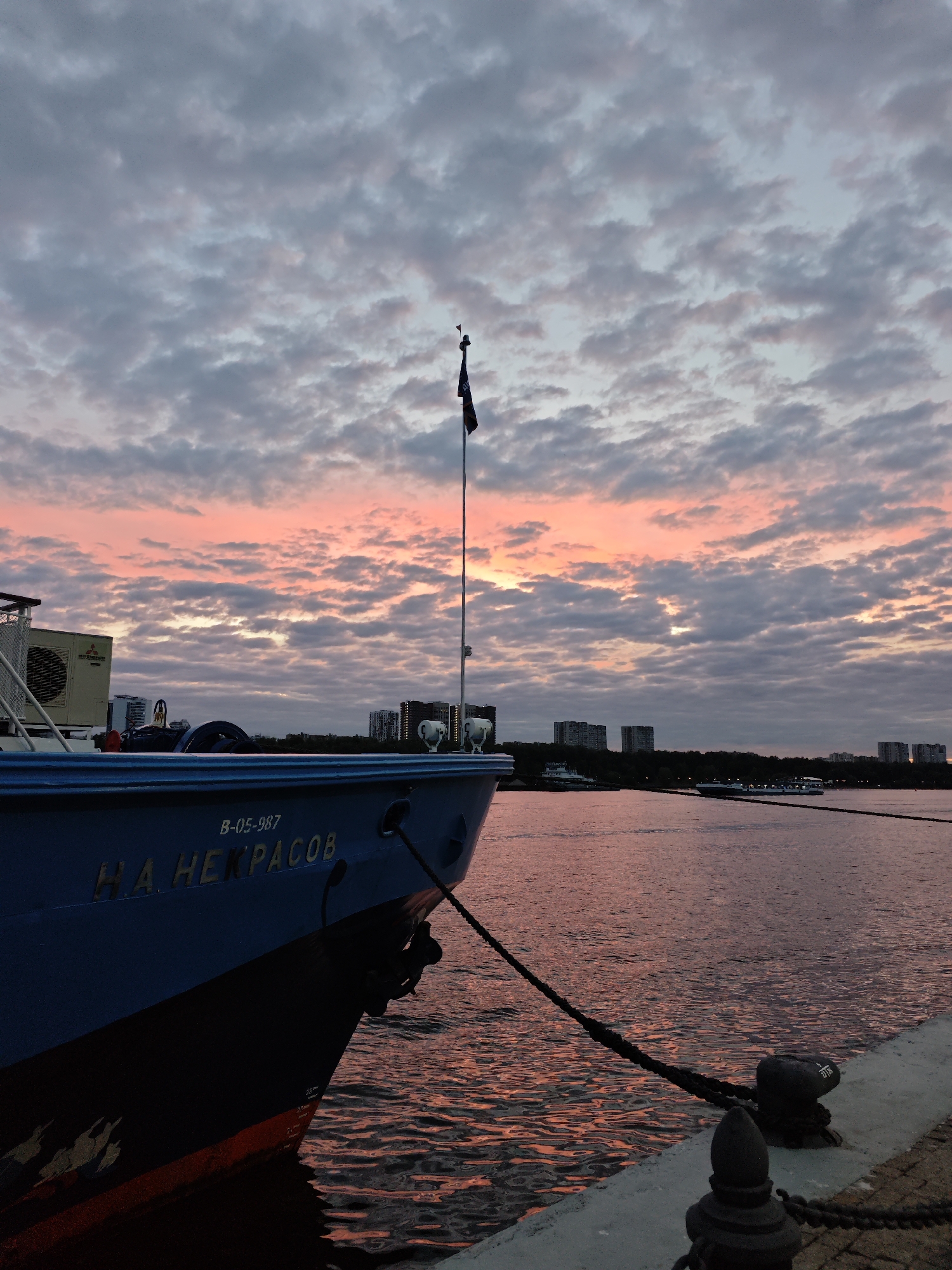 The image depicts a serene sunset scene over the Moskva River in Moscow, Russia. The foreground is dominated by the bow of a blue ship named "Н. А. НЕКРАСОВ" with the identification number "В-05-987" visible. The ship is moored at a concrete embankment, with a thick black rope secured to a bollard. An anchor is visible on the ship's bow. In the background, the river reflects the warm hues of the sunset sky, with scattered clouds tinged pink and orange. On the far bank, a line of residential buildings and trees are silhouetted against the horizon. A flag flies from a mast on the ship. The overall atmosphere is peaceful and tranquil, capturing the beauty of a Moscow sunset.