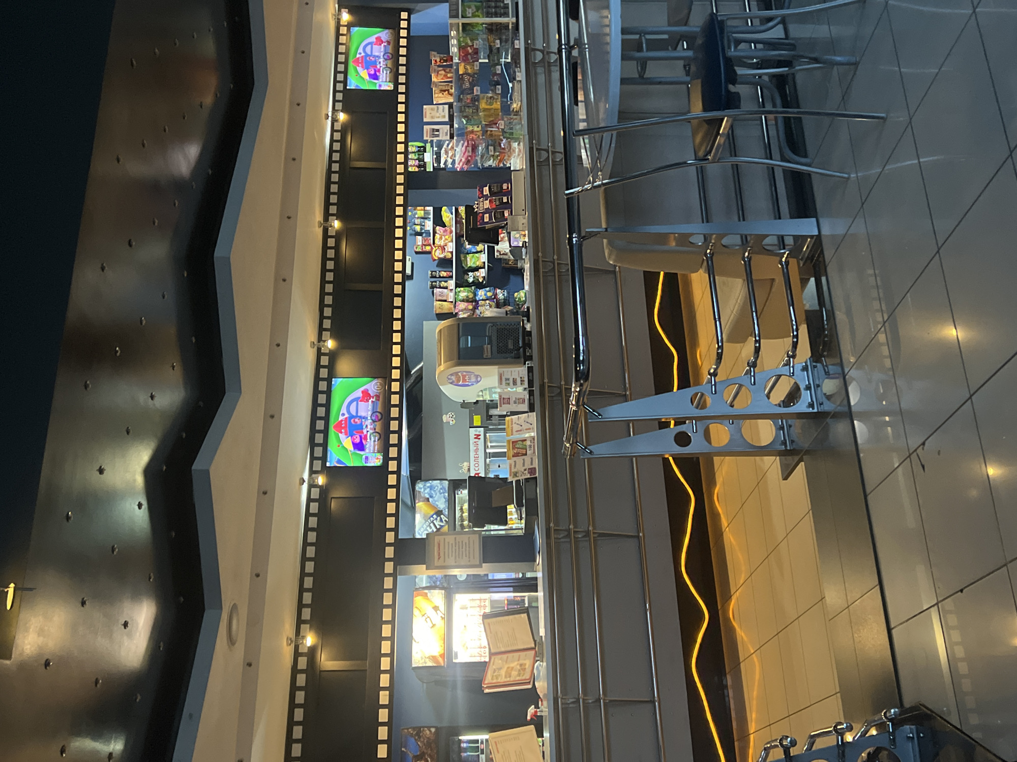 This media file captures an indoor scene at what appears to be a cinema or entertainment venue, possibly in Zheleznogorsk, Russia. The primary subject is a concession stand area, stocked with various snacks and beverages. The walls are decorated with film strip motifs, and screens display animated content, further suggesting a cinema setting.
The scene is well-lit, suggesting it is daytime or the venue is actively operating. The overall impression is one of a vibrant, commercial space designed for entertainment. There are no people clearly visible in the image, but the presence of seating and a counter implies that customers would typically be present.
Notable details include the variety of packaged snacks on display, the illuminated menu or promotional materials on the counter, and the architectural elements like the metal railings and the textured walls. The use of bright colors in the display screens and packaging adds to the lively atmosphere.
Some visible text includes what appears to be Russian language on the product packaging and signage. For example, "СОЛНЕЧНЫЙ" (Solnechny, meaning Sunny) is visible on a sign behind the counter. Other text is present on the snack packages and menus, but it is not fully legible. The overall ambiance is that of a typical concession area, designed to attract and serve patrons.