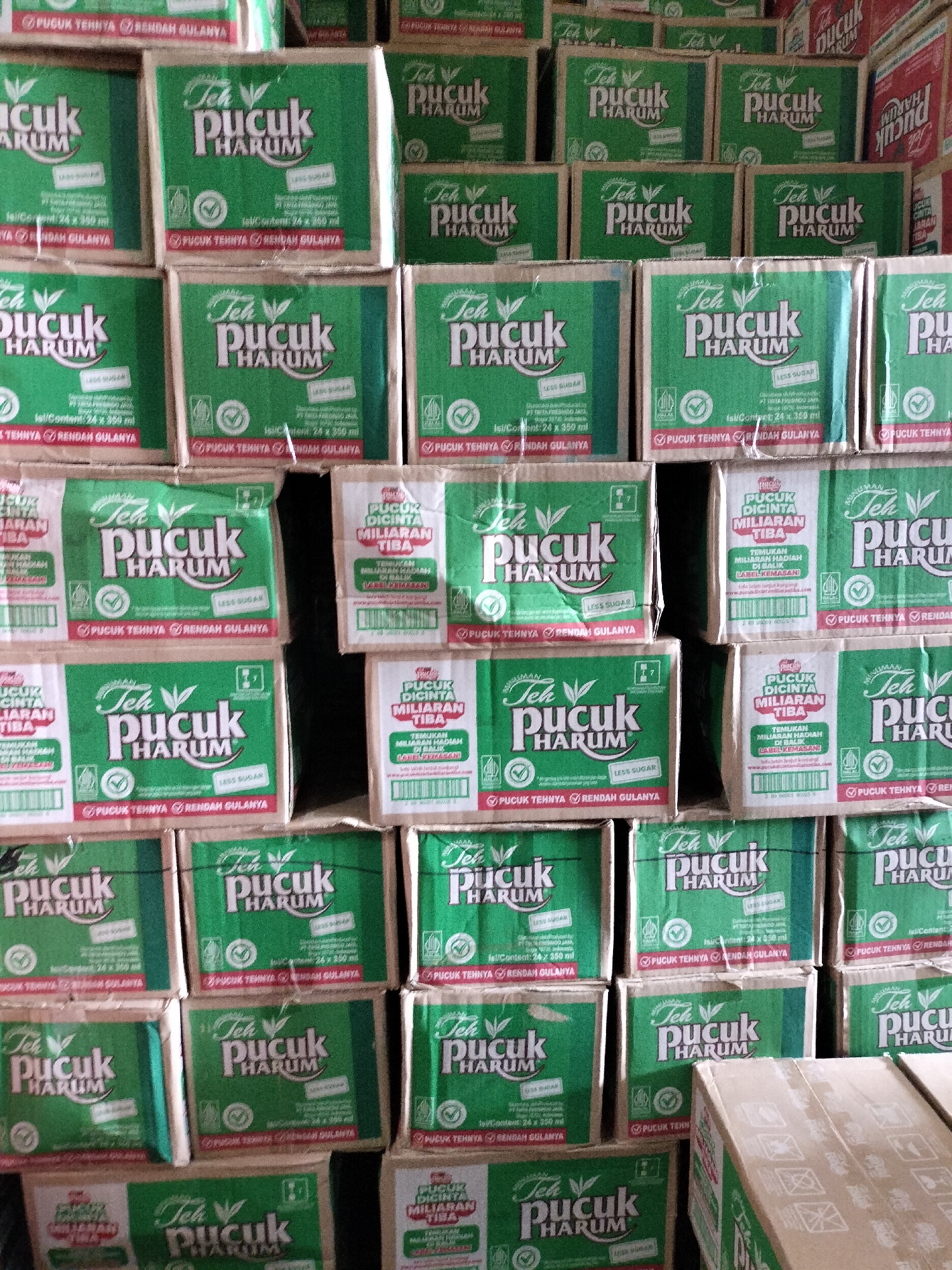 The image depicts a large stack of beverage boxes, primarily green and white, with the brand "Teh Pucuk Harum" prominently displayed. The boxes are arranged in rows and columns, indicating a retail or storage setting. The lighting suggests an indoor environment, possibly a warehouse or a shop. There are no people visible in the image. The boxes are likely tea products given the Indonesian text "Teh Pucuk Harum" which translates to "Jasmine Tea." The scene is static and offers no clues about specific activities or interactions. There is no information in the image that allows for the localization of this scene within the city of Langsa, Indonesia.