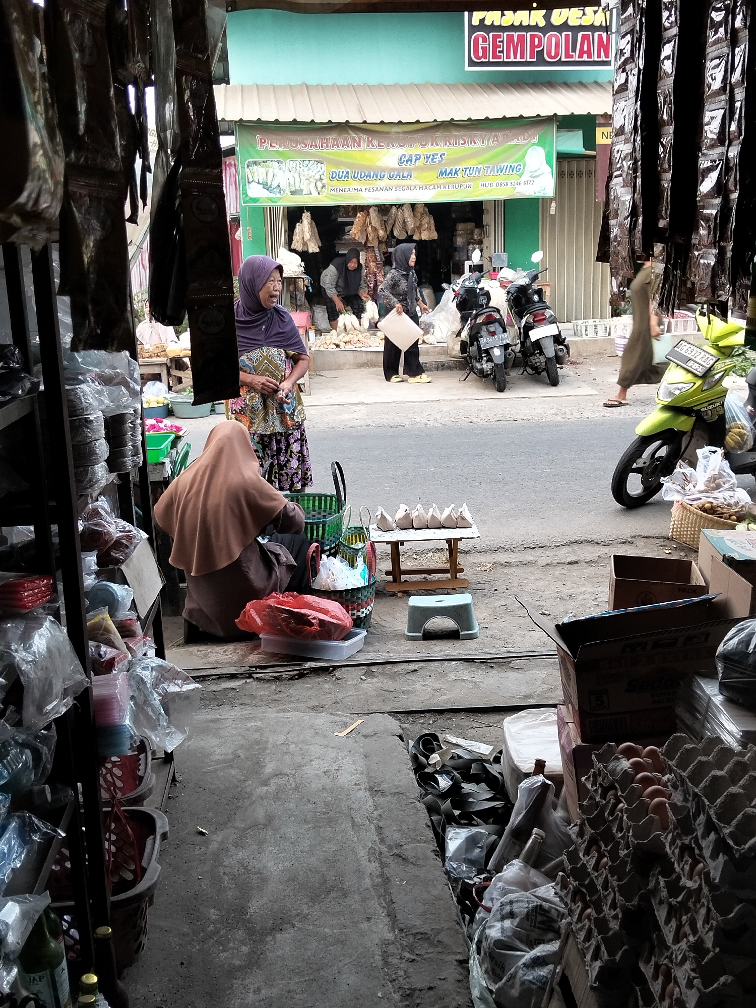 The image depicts a street market scene in Gempolan, Indonesia, identified by a sign above a store that reads "PASAR DESA GEMPOLAN." The foreground features a selection of goods for sale, including a prominent display of egg cartons filled with brown eggs, several cardboard boxes, and bags of various items. A person wearing a brown hijab is seated on the ground, facing away from the camera, with several shopping baskets and a red plastic container nearby. Behind this seated individual, another person wearing a purple hijab and a patterned top is standing and looking towards the left.
In the middle ground, a street is visible with several motorcycles parked. A bright green Suzuki motorcycle is prominently displayed on the right side of the street. Two women are interacting near the parked motorcycles; one is wearing a black hijab and a patterned top, and the other, partially obscured, is wearing a light-colored headscarf and holding a white board. A green banner with Indonesian text, including "DUA UDANG GALA" and "MAK TUN TAWING," is hanging from the storefront.
The background shows the storefront with its green awning and signs, indicating a commercial establishment. The overall environment is an outdoor market setting with a mix of goods, vehicles, and people engaged in commercial activity.