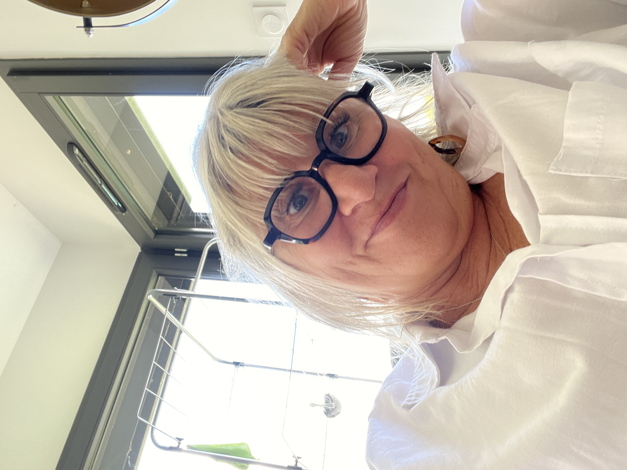 A close-up, slightly angled view shows a fair-skinned woman with short, light-blonde hair and dark-rimmed glasses. She wears a white collared shirt. The setting is indoors, with a window in the background, and what appears to be a clothes drying rack visible. Location is in the city of Chambœuf, France.