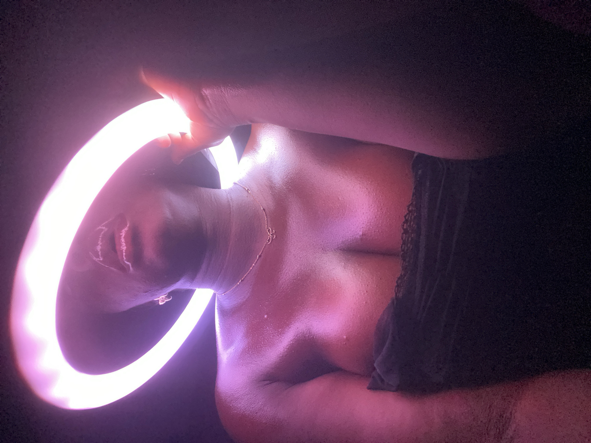 A person is lying down, oriented horizontally in the frame, and is strongly illuminated by a circular light source. The person's head is close to the left side of the image. A necklace is visible on the person's neck. The scene has dark surroundings, possibly inside a room. The location in the city of Maigizo, Nigeria, cannot be determined from the image.