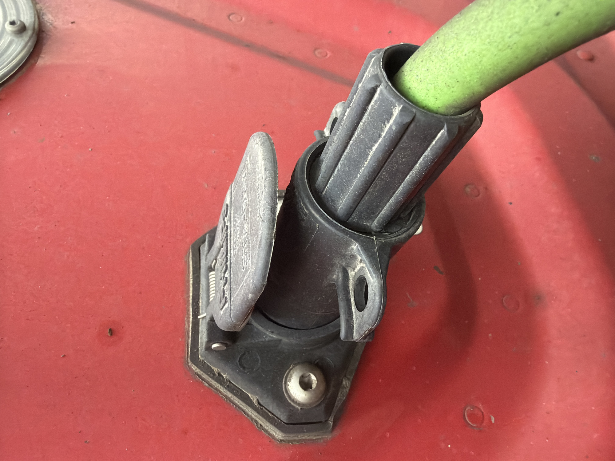 The image shows a close-up, eye-level view of a black electrical connector socket mounted on a red, textured surface, likely the exterior of a vehicle or piece of equipment. A green hose or cable is inserted into the connector. The socket is hexagonal with a hinged cover, which is partially open, revealing the internal contacts. A silver screw is visible on the left side of the socket, securing it to the red surface. The surface itself has several small, circular indentations and some dust or debris. The lighting suggests an outdoor environment, and the overall impression is one of functional, utilitarian equipment. There are no people or other distinguishing features in the image. The provided information does not specify a location in Murfreesboro, United States of America.