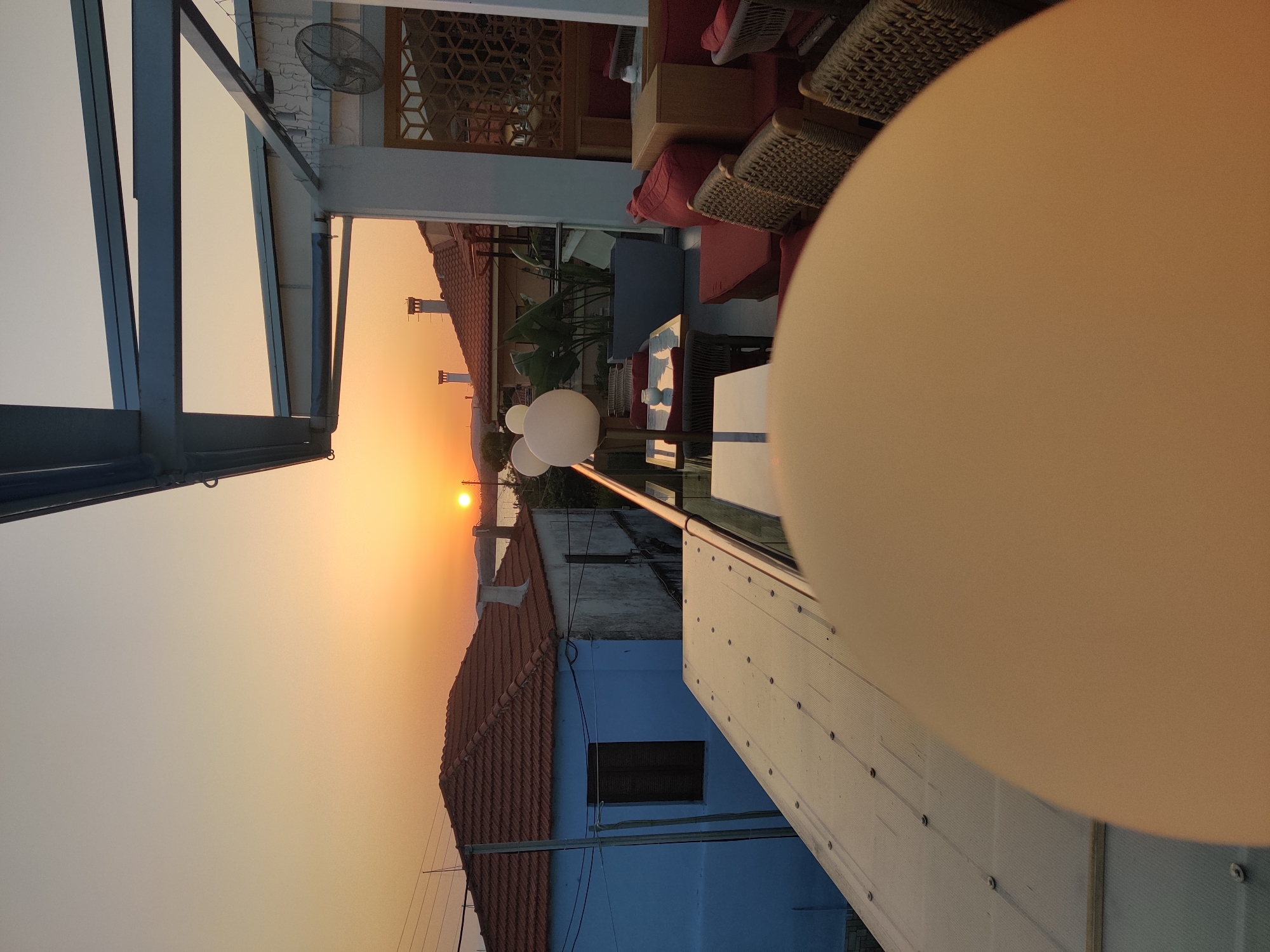 The image captures a sunset over a coastal town, likely in the Sithonia Municipal Unit of Greece, given the architectural style of the buildings with terracotta tiled roofs and the presence of the sea in the distance. In the foreground, a rooftop terrace area of a restaurant or cafe is visible. Several tables and chairs, some with woven wicker details and plush red cushions, are set up, suggesting an outdoor seating area. Decorative globe-shaped lights are also present. Beyond the terrace, traditional Greek houses with blue and white painted walls and tiled roofs are clustered together. The sky is awash with warm, orange and yellow hues of the setting sun, which is visible as a bright orb on the horizon. The overall scene conveys a peaceful and idyllic atmosphere, indicative of a Mediterranean evening.