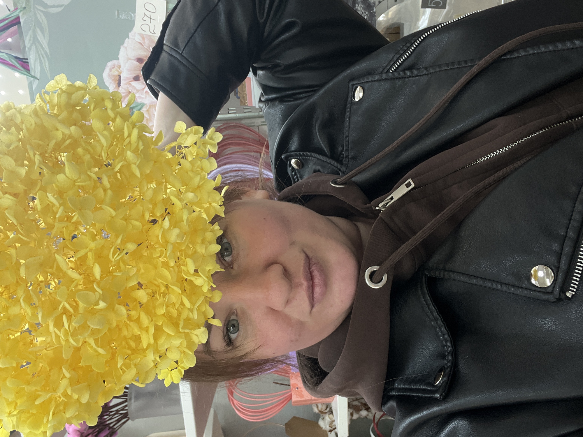 A female individual is depicted in a close-up, eye-level selfie taken indoors, likely within a retail establishment such as a flower or craft shop. She has light brown hair, green eyes, and a fair complexion with reddish cheeks, exhibiting a slight smirk. She wears a dark brown hooded sweatshirt beneath a black faux leather jacket featuring silver snap buttons and zippers. A large, spherical cluster of bright yellow flowers, consistent with dried or artificial hydrangeas, is positioned directly above her head, held in place by her right hand, which is visible at the upper right. The background includes white shelving units displaying various items: pink decorative objects (possibly heart-shaped wires), a grey vase containing pink artificial flowers, and cotton bolls. To the upper right, a light green surface with a subtle floral pattern is visible, affixed with a white label marked "270" in black numerals. The precise location within the city of Chaikovsky, Russia, cannot be determined from the image.