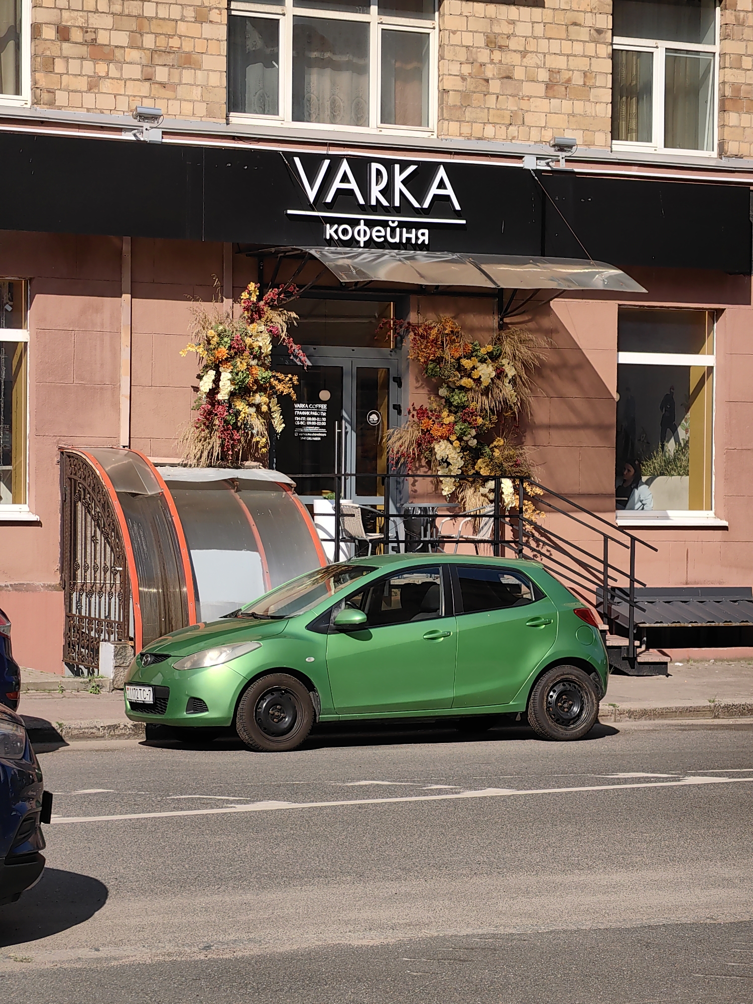 A green Mazda car is parked on the side of a city street in front of a building with a sign that reads "VARKA КОФЕЙНЯ". The building appears to be a cafe or coffee shop, as indicated by the word "кофейня" (coffee shop) in Russian. The facade of the building is made of reddish-brown brick, and it has large windows. There are also decorative floral arrangements on either side of the entrance. The street in front of the building is paved, and there is a sidewalk. The image is taken during the daytime, with sunlight casting shadows. The license plate of the car is partially visible and appears to be of Belarusian origin. The environment suggests an urban setting in Minsk, Belarus.