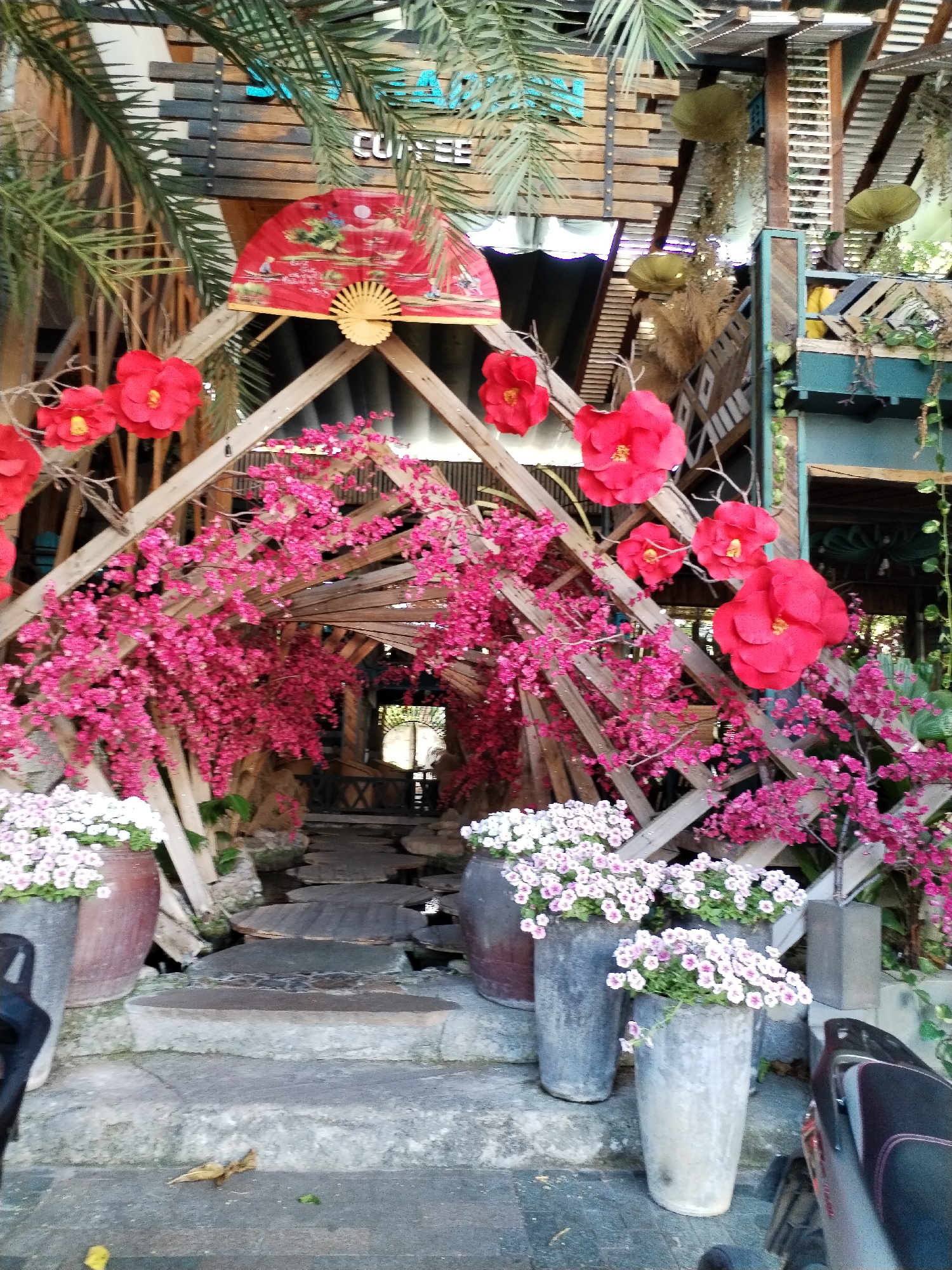 The image depicts a decorative entrance in Bac Nha Trang Ward, Vietnam. A wooden, A-frame structure is adorned with pink flowering vines and large, red paper flowers. A red fan is mounted at the apex of the structure. Potted plants with white and light pink flowers are positioned on the steps leading into the tunnel-like entrance. The flooring consists of irregular stone slabs. The surrounding architecture includes wooden structures and greenery. A motorcycle is partially visible on the right side of the frame.