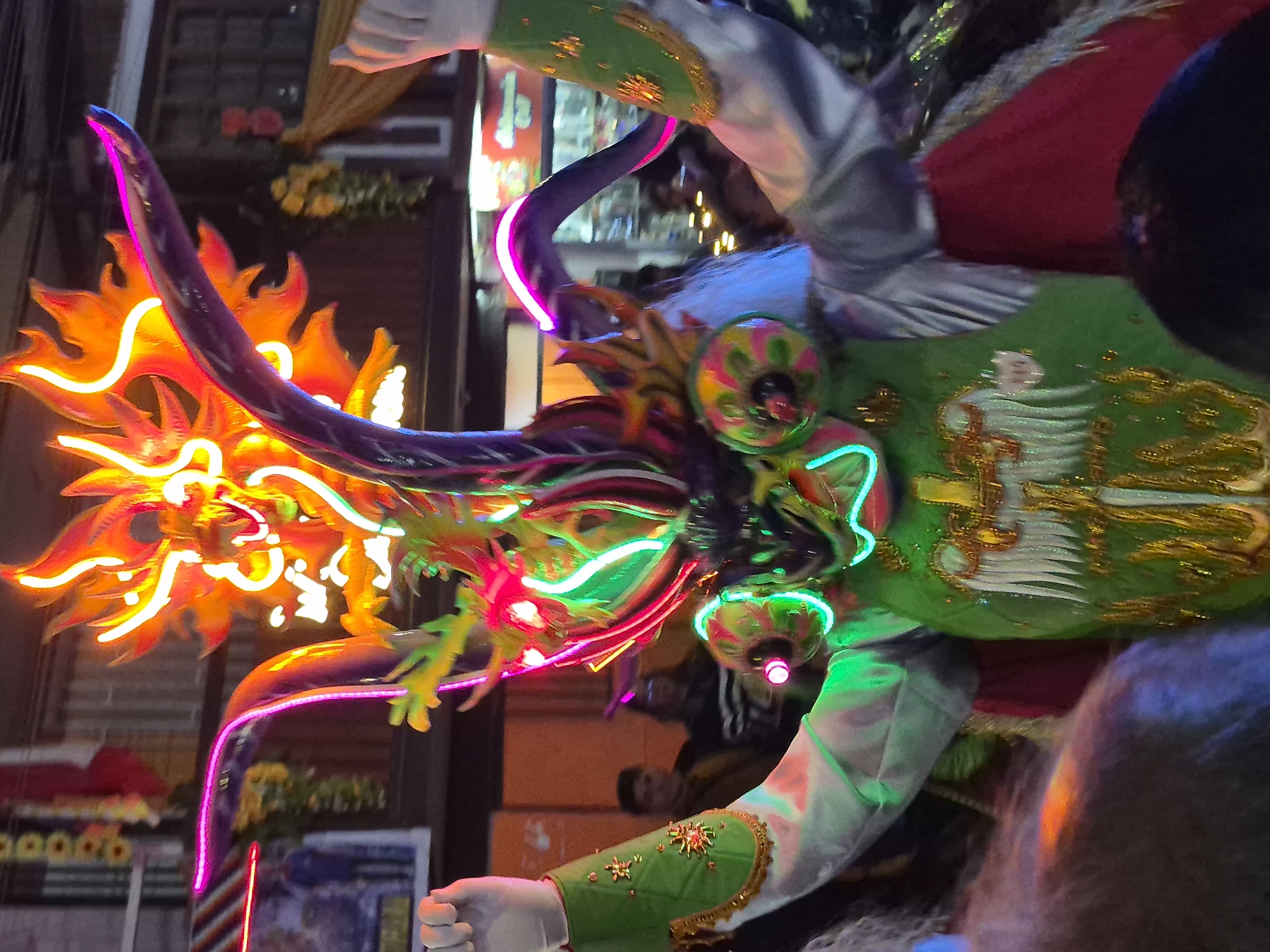 The image captures a vibrant and elaborate costume, likely from a festival or parade, in Pozo Almonte, Chile. The central figure is adorned with a detailed, brightly lit headdress resembling a dragon or mythical creature, illuminated by neon lights in orange, yellow, red, green, and purple. The costume features large, curved horns and a mask with prominent, circular eye features and a snarling mouth, also outlined in neon lights. The body of the costume is predominantly green, adorned with gold embroidery, and the sleeves are silver. The individual wearing the costume has their arms raised, with one hand clenched into a fist and the other extended upwards. In the background, blurred figures of onlookers and the facade of buildings are visible, suggesting an urban setting during an event. The overall impression is one of a lively cultural celebration.