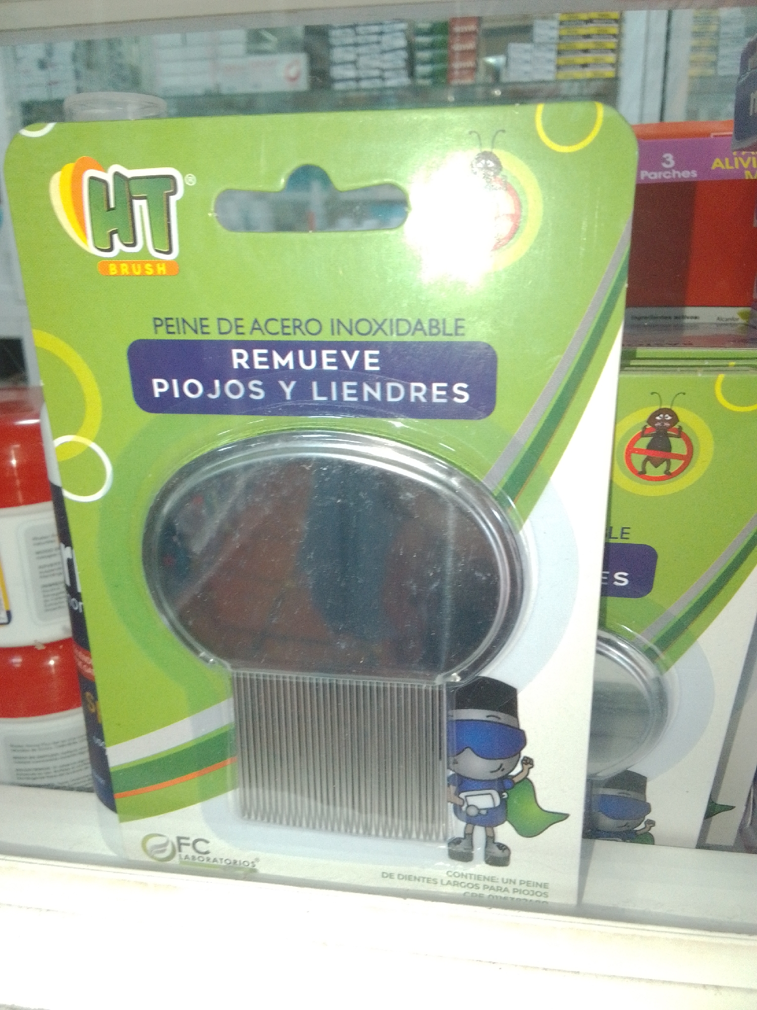 The image shows a retail display of a product labeled "HT BRUSH". The product is a stainless steel comb designed to remove lice and nits. The packaging is green and features cartoon illustrations of a ladybug and a character resembling a superhero with a cape. The product name and description are in Spanish. There are multiple units of the product displayed side-by-side. In the background, other retail products are visible on shelves. The lighting suggests an indoor retail environment, possibly a pharmacy or a general store. The image appears to have been taken at eye level, looking slightly upwards into the display. The location is identified as Juan Griego, Venezuela.