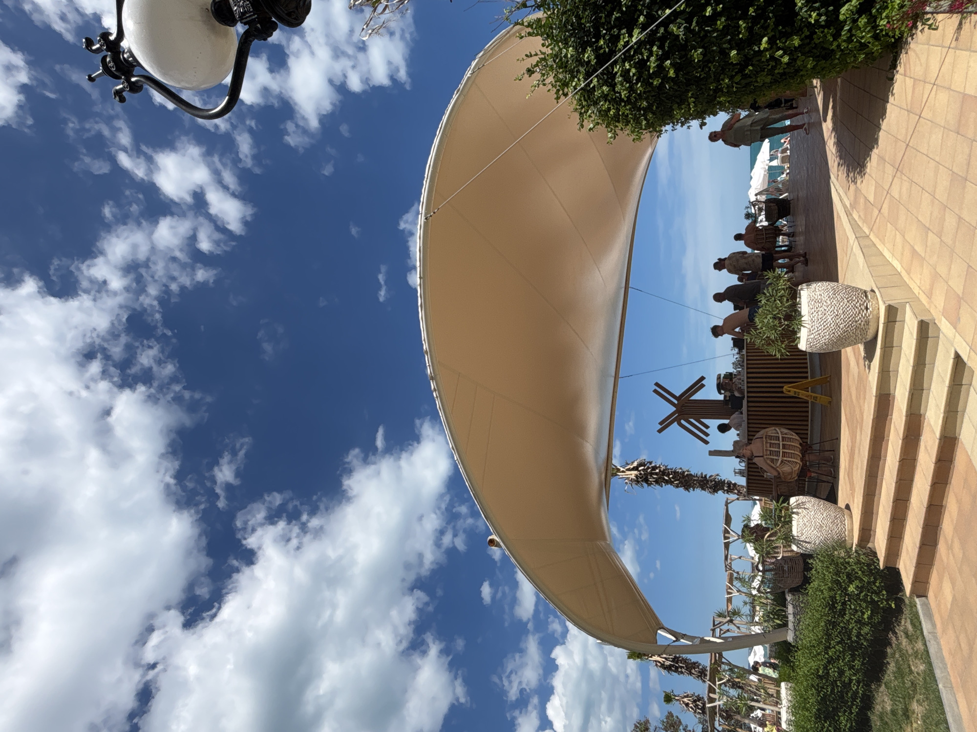 This is a wide-angle, eye-level shot of an outdoor beach bar on a sunny day. The sky is bright blue with scattered white clouds. A large, beige fabric canopy stretches across the frame, providing shade. In the foreground, there are tan tiled steps leading up to a seating area. To the left, there's a well-maintained green hedge and two large white planters, one with a green plant.
Under the canopy, a wooden bar is visible with several people standing and sitting. Some individuals are at the bar, while others are seated in wicker chairs. To the right of the bar, a woman in a green outfit stands, facing away from the camera, and a lush green bush is prominent.
In the background, beyond the bar area, a beach with white umbrellas and lounge chairs is visible, leading to the sea. Palm trees and other tropical-looking vegetation are interspersed throughout the scene. A vintage-style street lamp is visible in the upper right corner against the sky.
The overall impression is a relaxed, tropical resort atmosphere, likely located on a beach in Varna, Bulgaria, given the presence of outdoor bars and beach amenities.