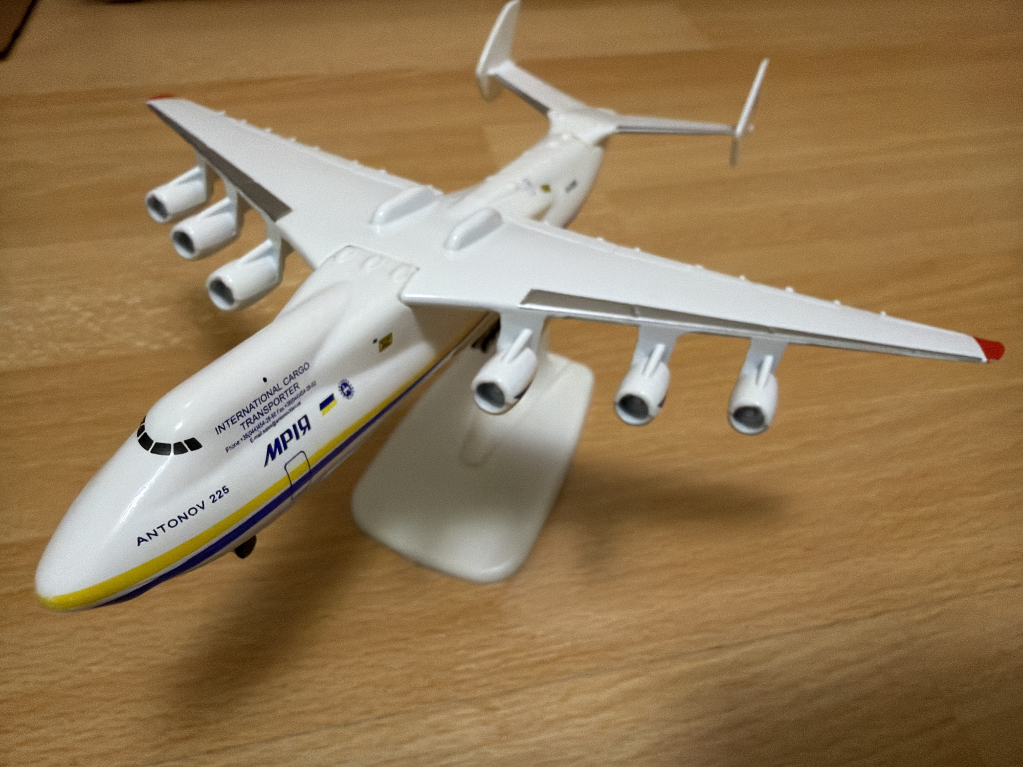 This is a close-up, high-angle shot of a white plastic model of an Antonov An-225 Mriya cargo plane. The model is positioned on a light brown wooden surface, likely a table. The plane is predominantly white with blue and yellow stripes along the fuselage. The text "ANTONOV 225" is visible on the front left of the fuselage, and "INTERNATIONAL CARGO TRANSPORTER" is printed above the Ukrainian flag colors and the designation "MРIЯ". The model has six engines mounted under its wings, three on each side. The scene is static and appears to be indoors. There is no indication of the city of Kyiv, Ukraine.