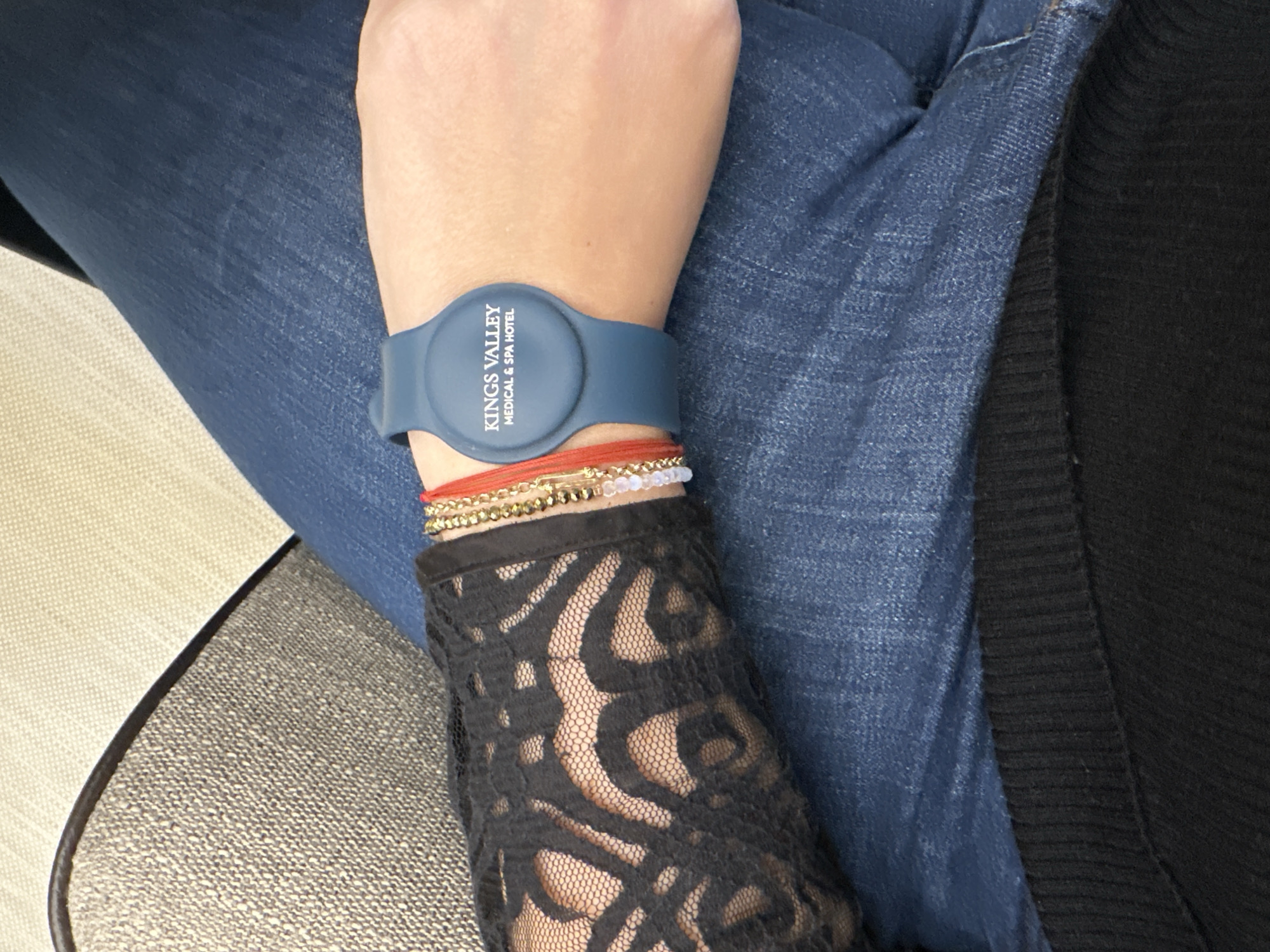 A close-up, high-angle shot captures a person's left arm and lap, highlighting a blue silicone wristband worn on the wrist. The wristband features white text arranged vertically, clearly reading "KINGS VALLEY MEDICAL & SPA HOTEL." Beneath the wristband, several other thin bracelets are visible, including a red string bracelet, a gold-beaded bracelet, and another with small white or clear iridescent beads.
The person's forearm is covered by a black lace sleeve, showcasing a patterned design. Their lap is covered by blue denim fabric, indicating they are wearing jeans. To the right, a section of black ribbed fabric is visible, likely part of another garment. On the lower left side of the frame, a textured beige or light brown upholstered surface, possibly a seat or armrest, with a dark trim, suggests an indoor setting. The scene depicts a static moment, focusing on the person's attire and accessories, particularly the branded wristband from what appears to be a medical and spa hotel, possibly located in Kazanlak, Bulgaria, given the provided context. No specific activity or emotion is discernible.