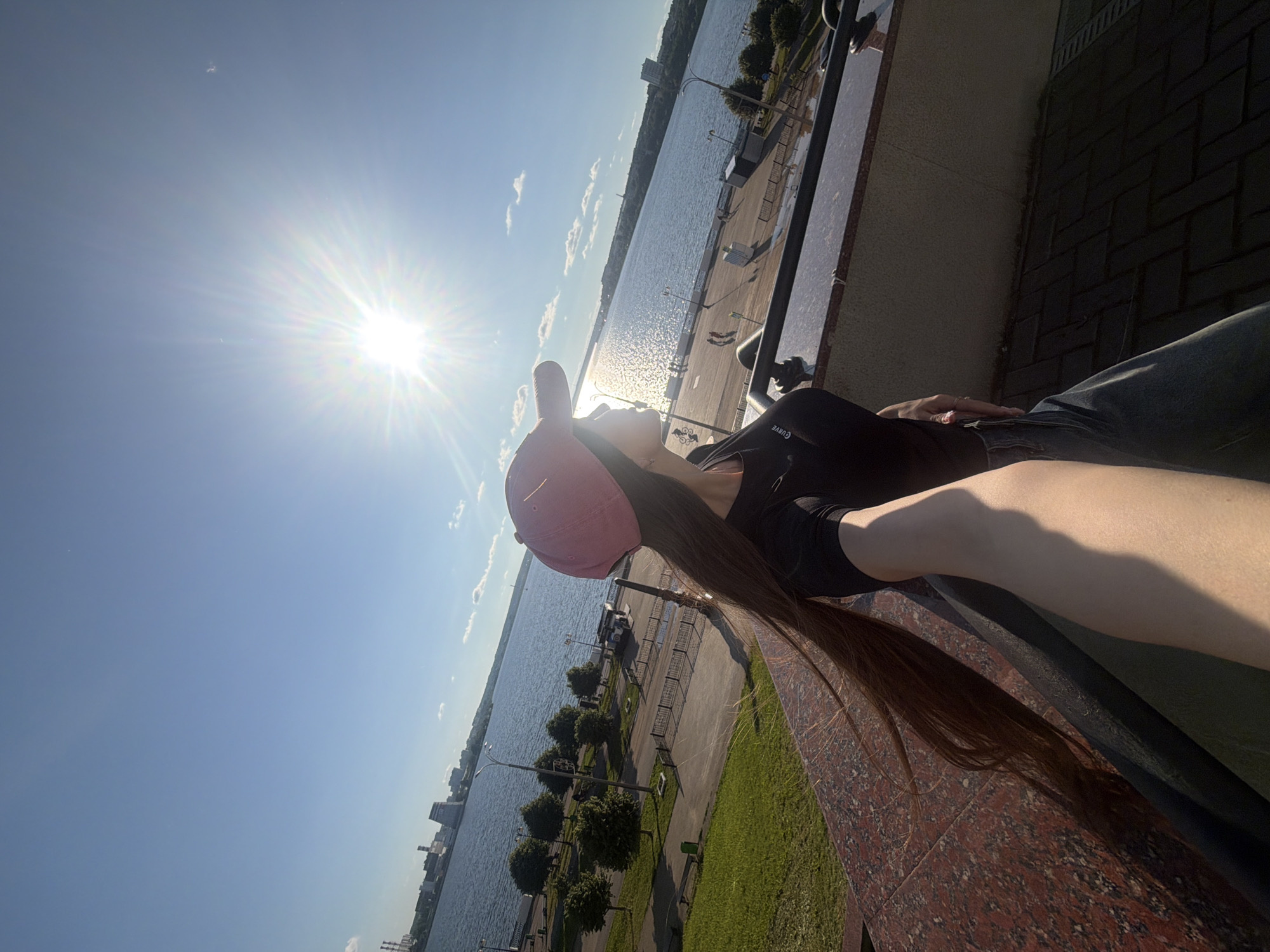 This media file captures a sunny outdoor scene in Izhevsk, Russia, featuring a person relaxing by a large body of water. The subject, a person with long dark hair, is seen from a side profile, wearing a pink baseball cap, a black top, and blue jeans. They appear to be sitting or leaning against a reddish-brown marble-patterned ledge or railing, with their head tilted slightly upwards.
The background showcases a wide expanse of water, likely a river or lake, possibly the Izhevsk Pond. Along the near bank, an embankment features a paved area, patches of green grass, small manicured trees, and streetlights. Buildings and a distant shoreline are visible across the water.
A prominent, intensely bright sun is positioned in the upper left of the frame, creating strong sunbeams and lens flare, indicating a clear, very sunny day, likely midday or early afternoon. The sky is a vibrant blue with minimal cloud cover. The person's pose suggests a moment of relaxation, possibly looking out at the water or towards the sun. The word "CURVE" is partially visible on the black top worn by the subject.