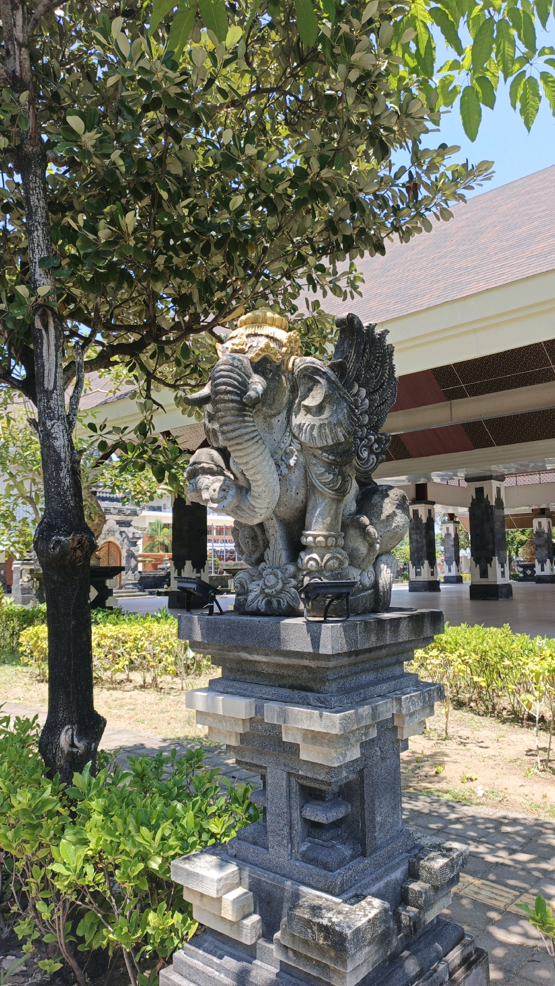 A stone sculpture of a winged elephant is positioned outdoors in front of a building with a reddish-brown tiled roof and white pillars. The sculpture is ornately carved, with a golden headdress and detailed wings. It rests on a multi-tiered stone pedestal. Lush green foliage, including a tree with a thick trunk to the left and bushes in the foreground and background, surrounds the sculpture. The ground is paved with light-colored stones, and some patches of dry earth are visible. The sky is clear and blue. The overall setting appears to be a temple or cultural site in Denpasar, Indonesia, characterized by traditional Balinese architecture and landscaping. There are no people visible in the image.