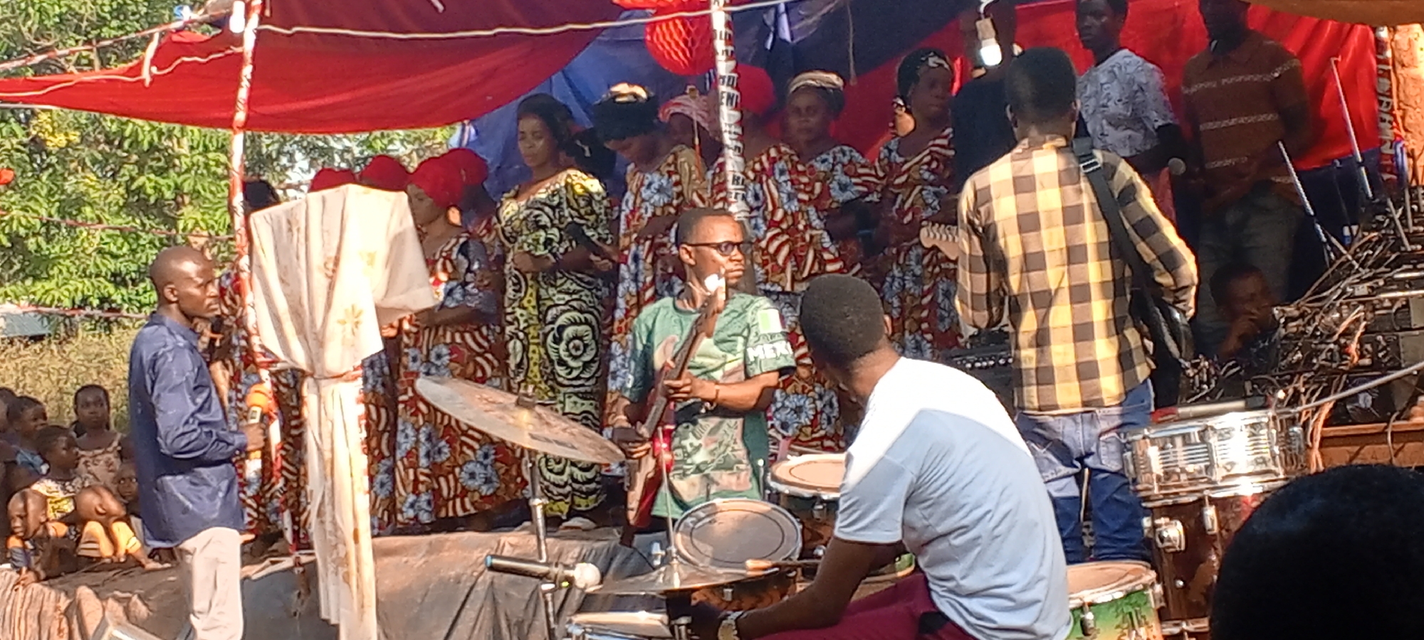 Captured in Ngalula, Democratic Republic of the Congo, the image presents a lively performance scene. A group of individuals are gathered on a raised stage or platform. Several women, dressed in patterned, colorful traditional clothing, stand in the background, possibly forming a choir or ensemble. Two men are actively engaged in playing musical instruments. One is seated behind a drum set, and another standing beside him playing a bass guitar. A man is standing to the left with a microphone in hand. An individual in a checkered shirt with a strap over his shoulder stands in the foreground. A number of children are seated or gathered to the left of the stage. The stage is partially covered by a canopy of red and blue fabric. A few decorative elements, such as what appears to be a red paper lantern, are visible. Audio equipment including wires, speakers, and amplifiers, can be seen stage right.