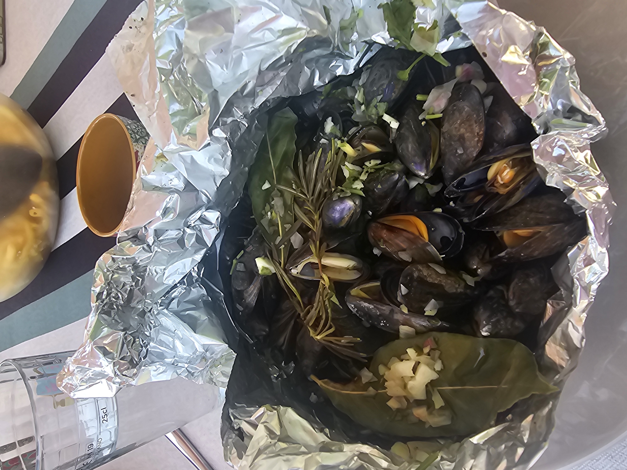 A close-up, overhead shot captures a meal of cooked mussels served in a foil packet on a table with a striped tablecloth. To the left, a glass of water is visible, filled to the 25cl mark. In the background, a yellow bowl contains pasta. The foil packet, a dark iridescent color, is open, revealing the mussels interspersed with herbs and what appears to be chopped garlic and onion. Some mussels are open, showing their orange interiors. The overall lighting is bright, suggesting an outdoor or well-lit indoor setting. There are no people or explicit signs of location, however, the presence of mussels, often prepared in such a manner in coastal regions, could suggest a seaside location. The exact location in Rozès, France cannot be determined from the image alone.
