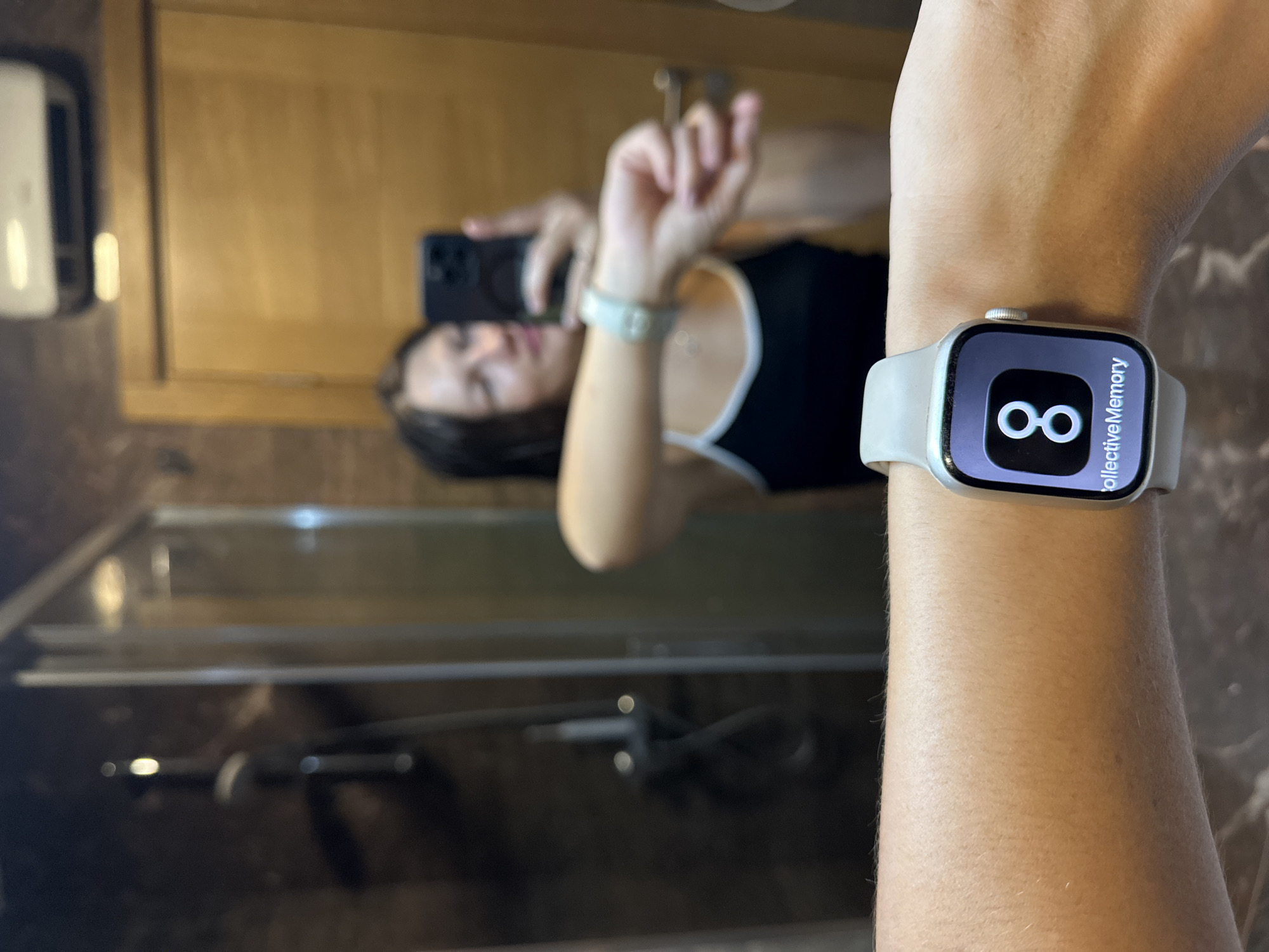 The image shows a person's arm with a smartwatch displaying the number "8" and the text "collectiveMemory". The person is in a bathroom, with a shower visible in the background and a wooden door reflected in the mirror. The location is identified as Lapčići, Montenegro. The person is holding a smartphone, taking a selfie. The smartwatch is light grey with a white band.