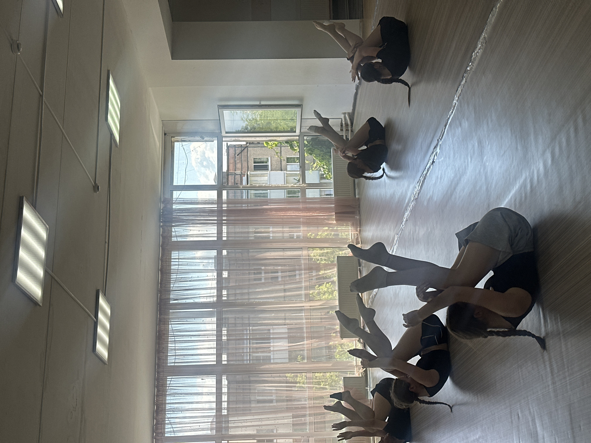A group of girls in black athletic wear are performing synchronized floor exercises in a dance studio. The studio has a wooden floor, large windows covered by sheer curtains, and fluorescent lighting fixtures on the ceiling. The girls are lying on their backs with their knees bent and feet lifted, some are holding onto each other's feet, suggesting a group choreography. The scene is illuminated by natural light coming through the windows and the overhead lights. The view outside the windows shows buildings and trees, indicative of an urban setting. The location is identified as Votkinsk, Russia.