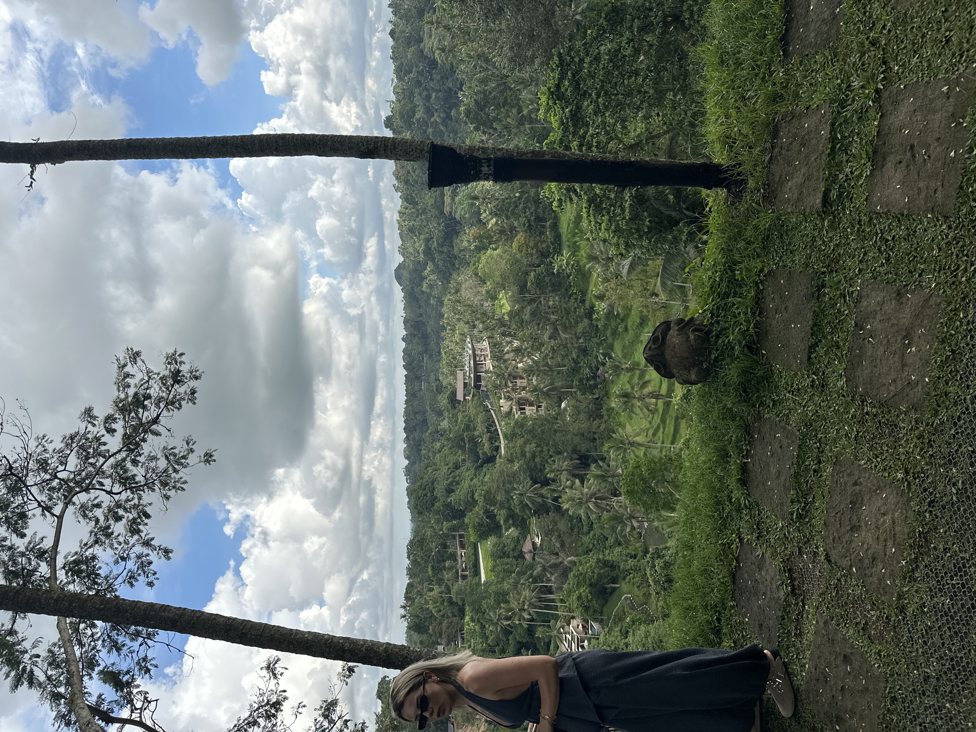 The image captures a scene in the Ubud District of Indonesia. A woman with blonde hair and sunglasses is leaning forward at the edge of an elevated walkway. She is wearing a dark gray dress.
The walkway is constructed of large, irregularly-shaped pavers with grass and vegetation growing between them. Adjacent to the walkway is a stone sculpture on the ground.
A thick, dark tree trunk spans the image horizontally. In the distance, a dense, green rainforest fills the frame, with glimpses of buildings and rice paddies. Above the forest canopy, a cloudy sky is visible.