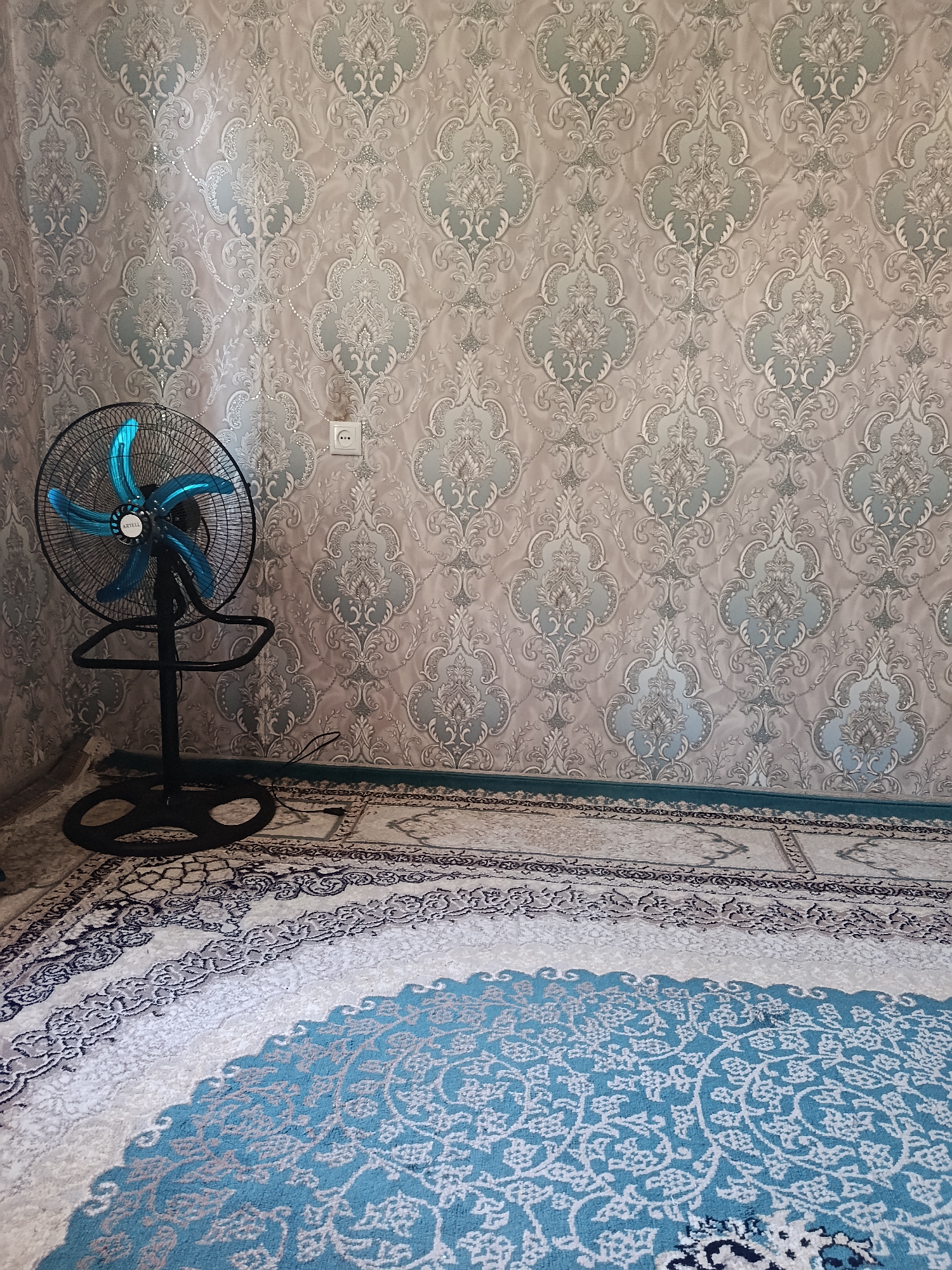 A black standing fan with bright blue blades is positioned to the left of the frame, in front of a wall covered in ornate, light-colored wallpaper. The wallpaper features a repeating damask pattern with a subtle sheen. To the right of the fan, a single white electrical outlet with three holes is visible on the wall. The floor is covered with a large rug, predominantly in shades of white and teal, with intricate floral patterns and a decorative border. The bottom edge of the wall meets the rug with a narrow band of teal trim. The image appears to be taken indoors, likely in a residential setting within the city of Gulbog 1, Tajikistan.