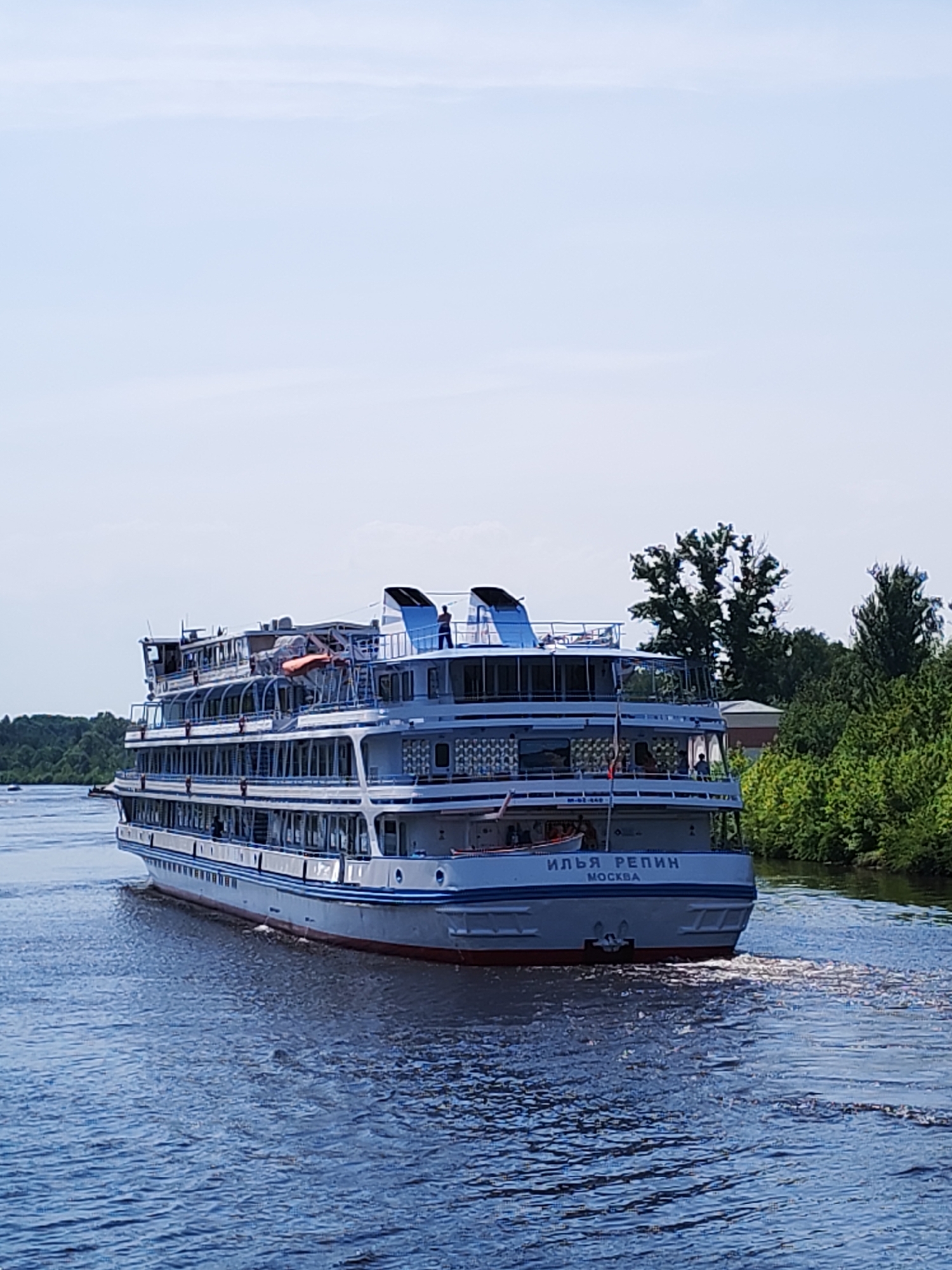 A medium shot captures a white cruise ship traveling through a waterway. The name "ИЛЬЯ РЕПИН" and "МОСКВА" are inscribed on the ship's stern. A solitary figure stands on the top deck. Trees line the right bank of the waterway, while more distant foliage is visible on the left.