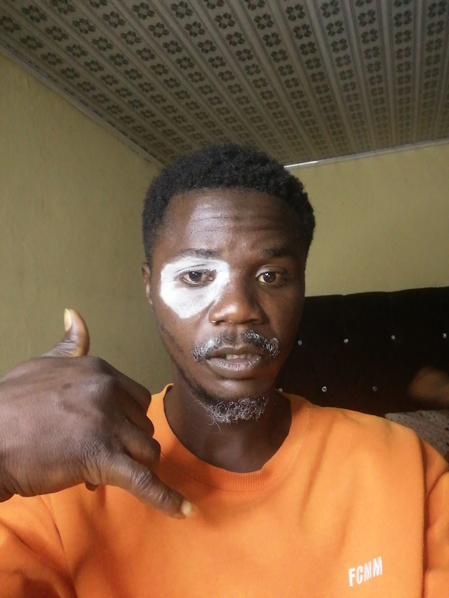 A dark-skinned man is featured in a close-up, selfie-style indoor photograph, presumably in Gura Top, Nigeria. He has short, dark, curly hair and a goatee and mustache that show visible grey hairs. A distinct white substance is applied around his left eye (viewer's left), extending from his eyebrow to his upper cheekbone.
The man is wearing a plain orange long-sleeved shirt or sweatshirt. On the lower right side of his shirt, the white letters "FCMM" are visible. He is making a "call me" hand gesture with his left hand (viewer's right), with his thumb and pinky finger extended and the other three fingers curled inward. His gaze is directed straight at the camera, and he has a neutral to slightly serious expression with his mouth slightly open.
The background shows a light yellow wall on the left side of the frame. To the right, a dark headboard or piece of furniture with some shiny elements, possibly studs, is partially visible. Above, a patterned ceiling with rows of repeating square-like motifs provides a distinctive architectural detail. The lighting indicates an indoor setting, with no specific time of day or weather discernible. The white facial marking and the prominent hand gesture are notable elements of the image.