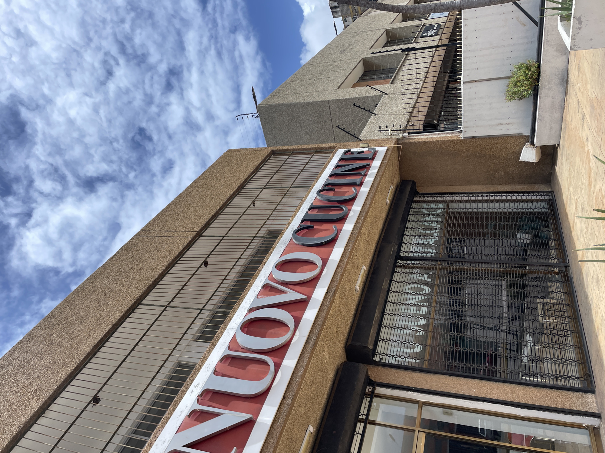 The image shows the exterior of a building with a sign that reads "NUOVO CUCINE". The building has a textured brown facade and a second story with barred windows. Below the sign, a storefront is visible behind a metal security gate. The storefront appears to be closed. In the background, another building with a similar facade and barred windows is visible. The sky is blue with some scattered clouds. The location is identified as Porlamar, Venezuela. The scene is one of an urban commercial area under daylight.