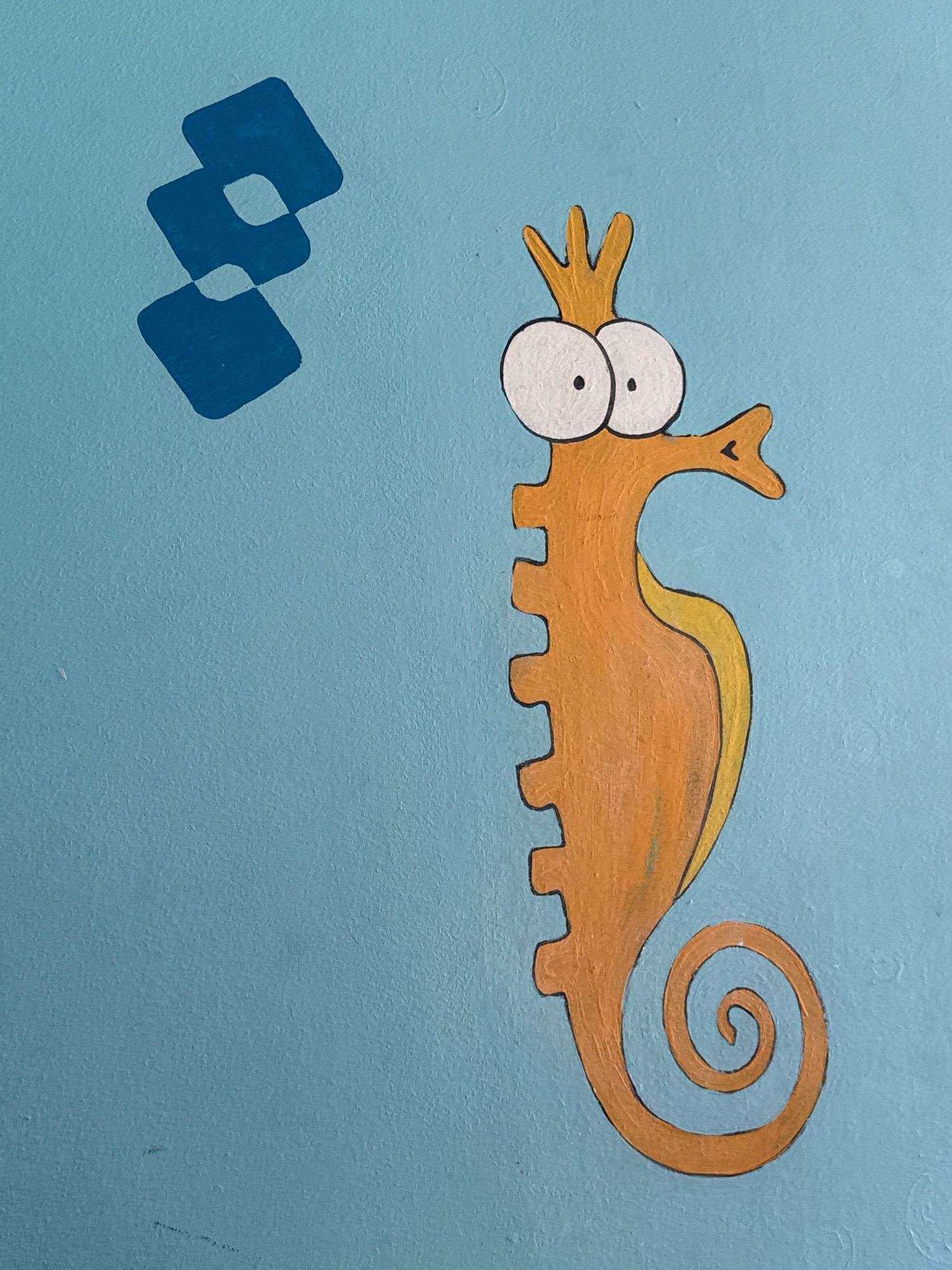 This is a close-up, eye-level, slightly angled shot of a cartoonish, orange seahorse painted on a textured light blue wall. The seahorse is depicted vertically, facing right, with large, wide white eyes with black pupils, and a crown-like protrusion on its head. Its body is segmented and it has a curled tail. To the left of the seahorse, there are three overlapping blue diamond shapes. The overall style is simple and childlike.
This image was taken in Sharm El Sheikh, Egypt.