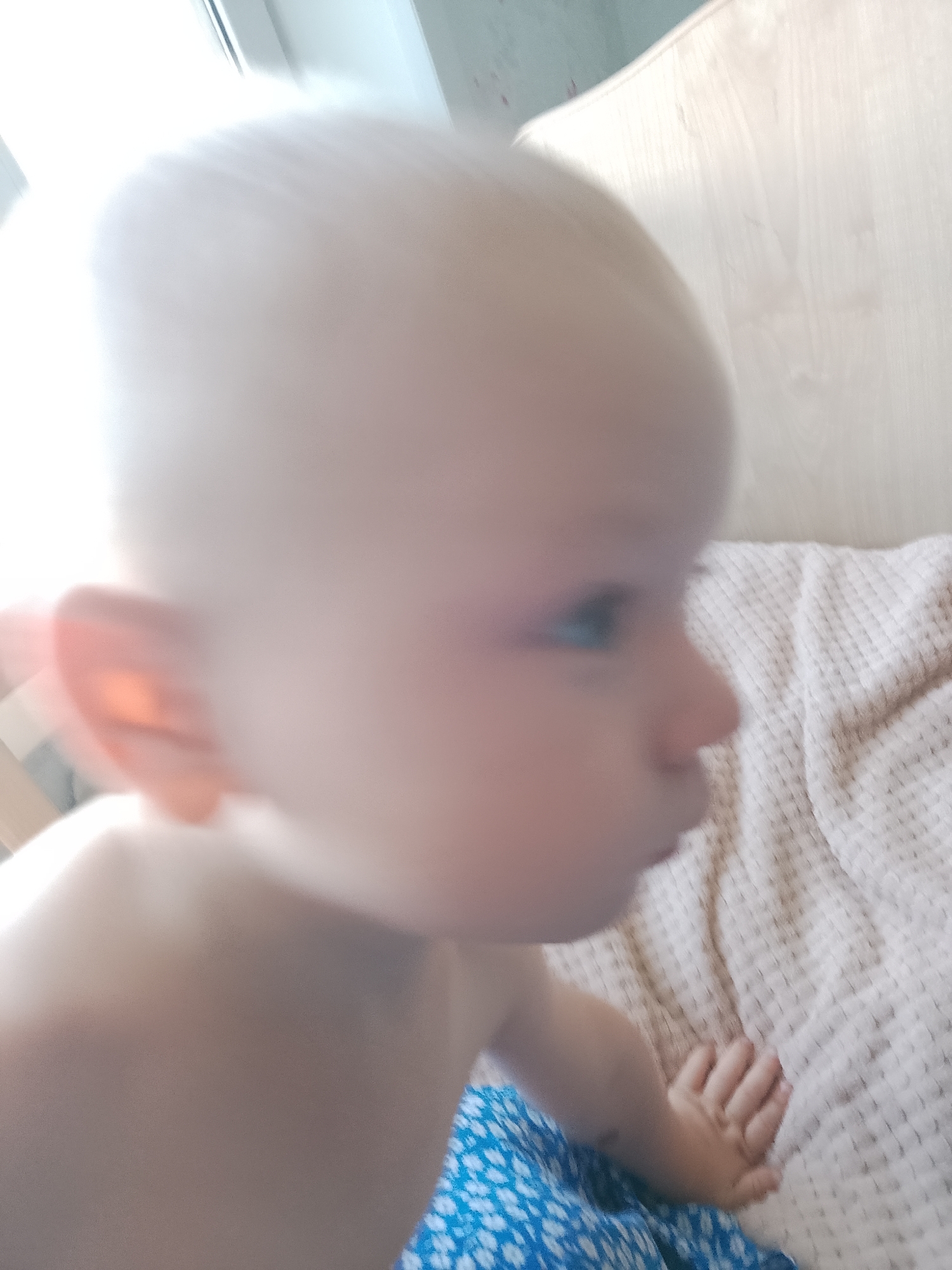 A very blurry, close-up, eye-level shot features a baby's head and shoulder. The baby has fair skin and light hair, and is facing to the right. The baby's right ear is visible, and the right eye appears to be blue. The baby is wearing a blue and white patterned garment. The background consists of a light-colored, textured surface, possibly a blanket or upholstered furniture, and a lighter area, possibly a window or bright light source. The overall impression is of a snapshot taken in a domestic setting. There is no information about the location within the city of Abay, Kazakhstan.