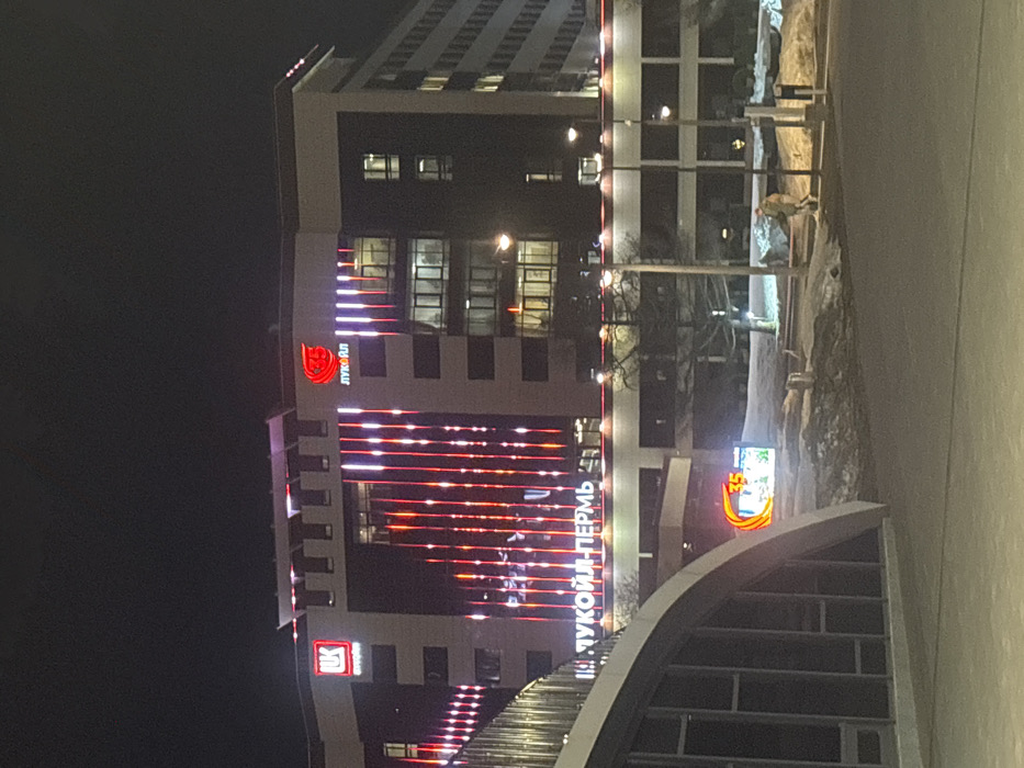 The image displays a nighttime scene in Perm, Russia, featuring the Lukoil-Perm office building. The multi-story structure exhibits a dark facade illuminated by internal window lights and external decorative red and white vertical strip lighting. Prominent red corporate signage includes the Lukoil stylized flame logo and 'ЛУКОЙЛ' text on the upper facade, with 'ЛК' and 'ЛУКОЙЛ-ПЕРМЬ' signs positioned on a lower section of the building's exterior. In the immediate foreground on the left, a curved architectural element composed of concrete and glass, indicative of a pedestrian overpass or underpass entrance, partially obstructs the view. The ground below is covered with snow, indicating cold weather conditions. Multiple streetlights cast illumination across the scene. A digital billboard displaying the number '35' is visible at ground level on the right. One individual, dressed in light outerwear, is observed walking on the snow-covered terrain in the mid-ground, positioned near the streetlights.