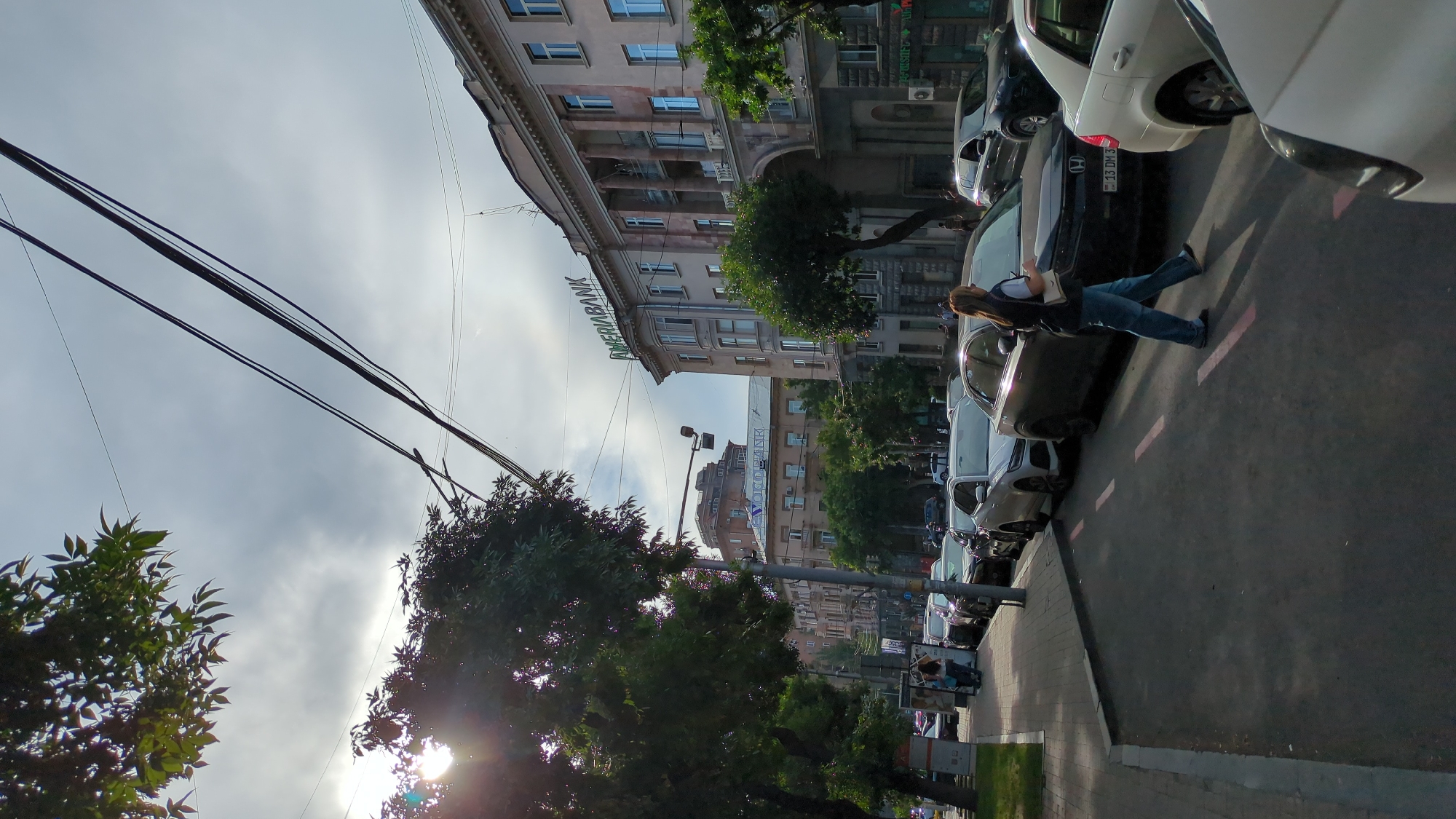 In Yerevan, Armenia, a street scene unfolds on a cloudy day with the sun breaking through the clouds, casting lens flare over a tree on the left. The street is lined with parked cars and a sidewalk. A woman with blonde hair, wearing a dark vest, white shirt, and blue jeans, walks across the street from right to left. A modern white car is parked to her right, and a dark sedan is parked to her left. In the background, several multi-story buildings are visible, including a building with "AMERIABANK" prominently displayed on its facade. Trees line the street and sidewalk, and overhead power lines are strung across the sky.